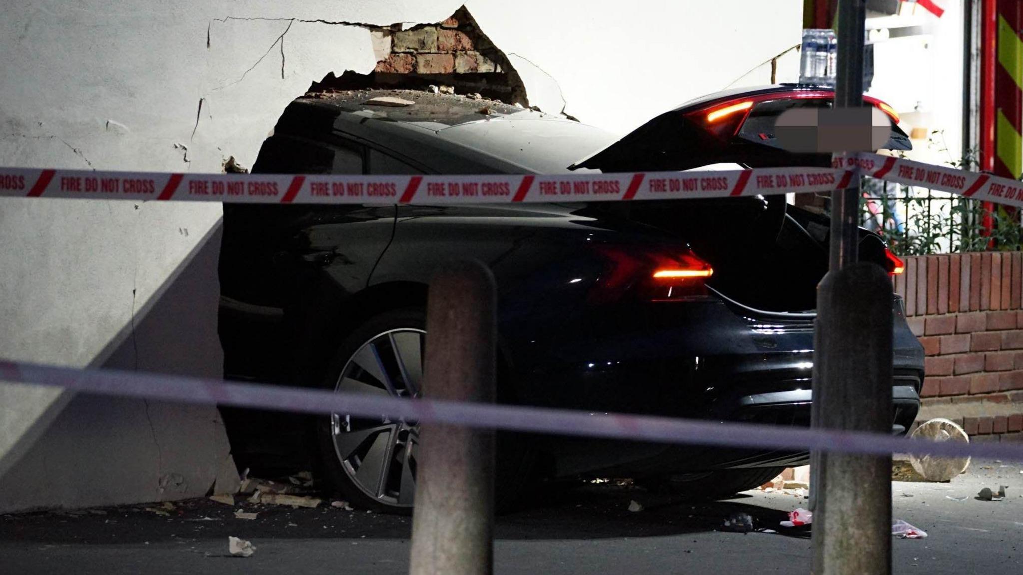 The end of a black car is visible with the rest having smashed through a white wall. 