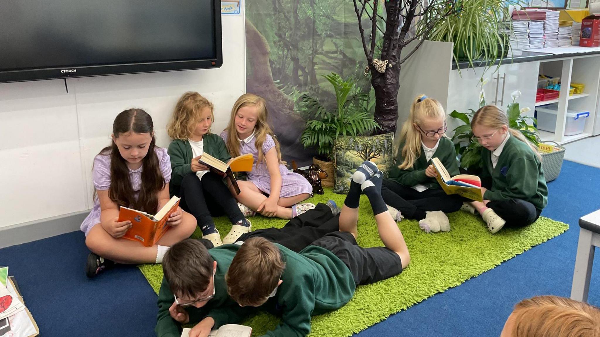 Seven children are reading. Two boys are lying on the floor, with a single girl to their right. Behind them are two pairs of girls sharing a book. They are on a bright green carpet and sitting at the base of a plastic tree.
