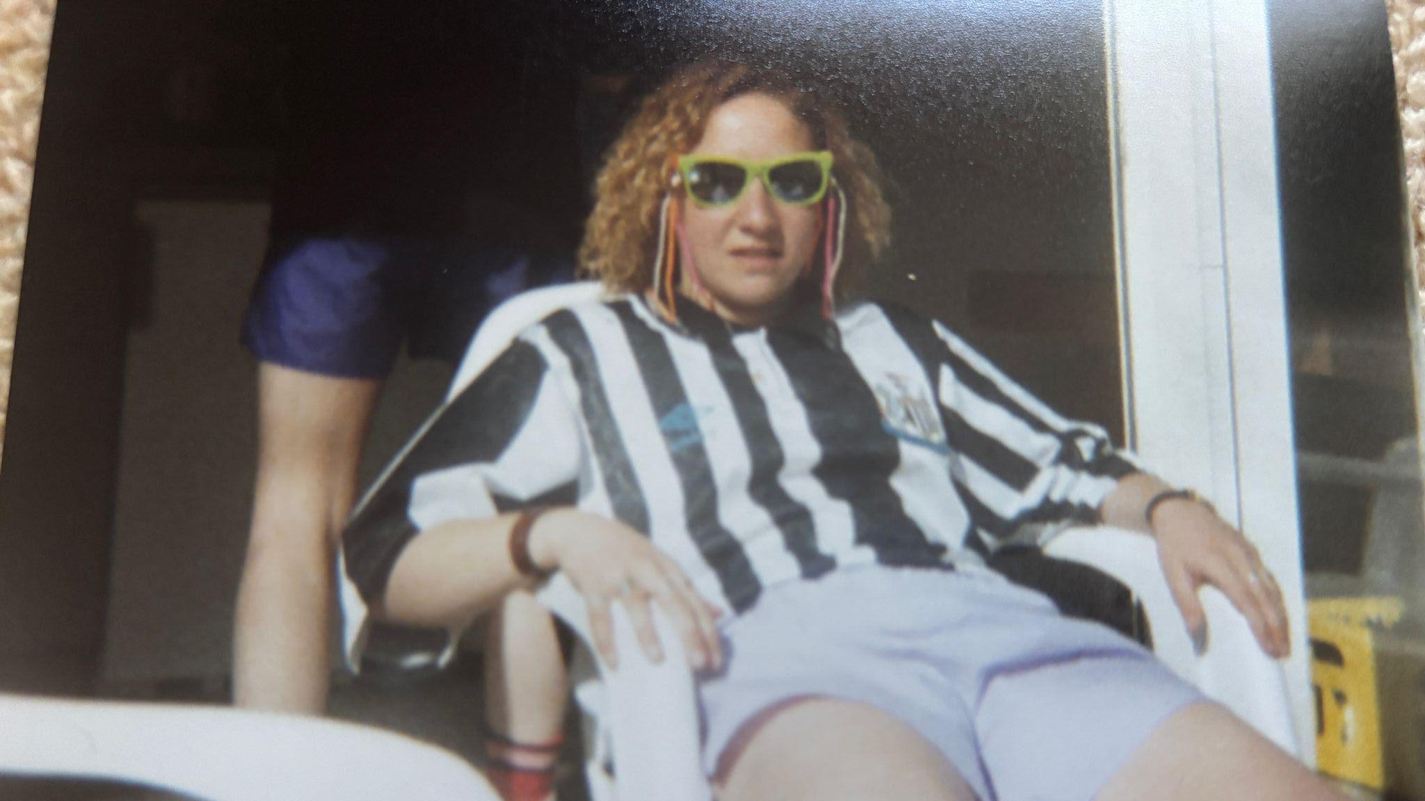 Lynn Simmonette with blonde curly hair, wearing yellow rimmed sunglasses and a black and white Newcastle football shirt, and grey shorts, sitting on a chair outside a patio door in the sunshine.