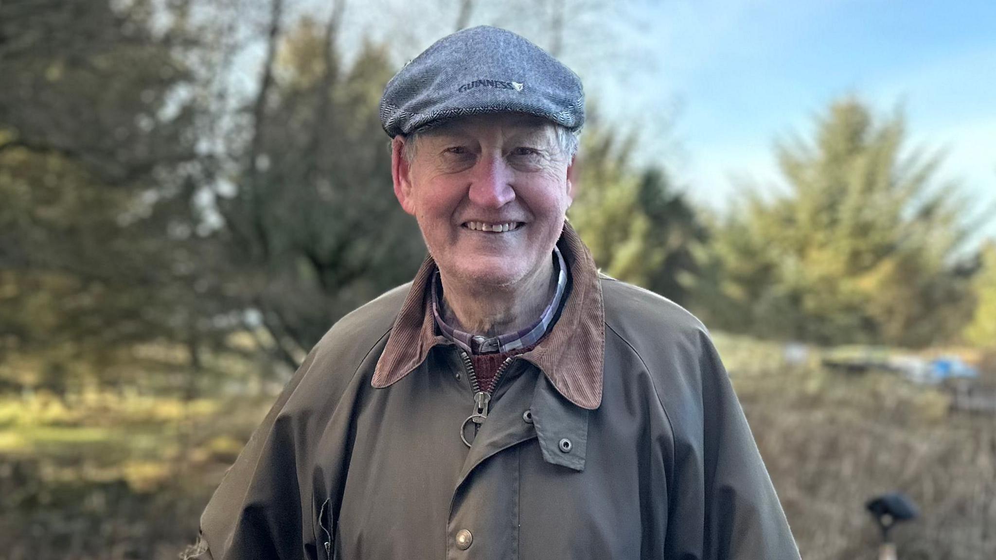 Patrick is pictured smiling in front of the trees and nettles. He is wearing a dark grey flat cap with 'Guinness' written on, along with a wax green shirt, a checked shirt and round neck jumper.