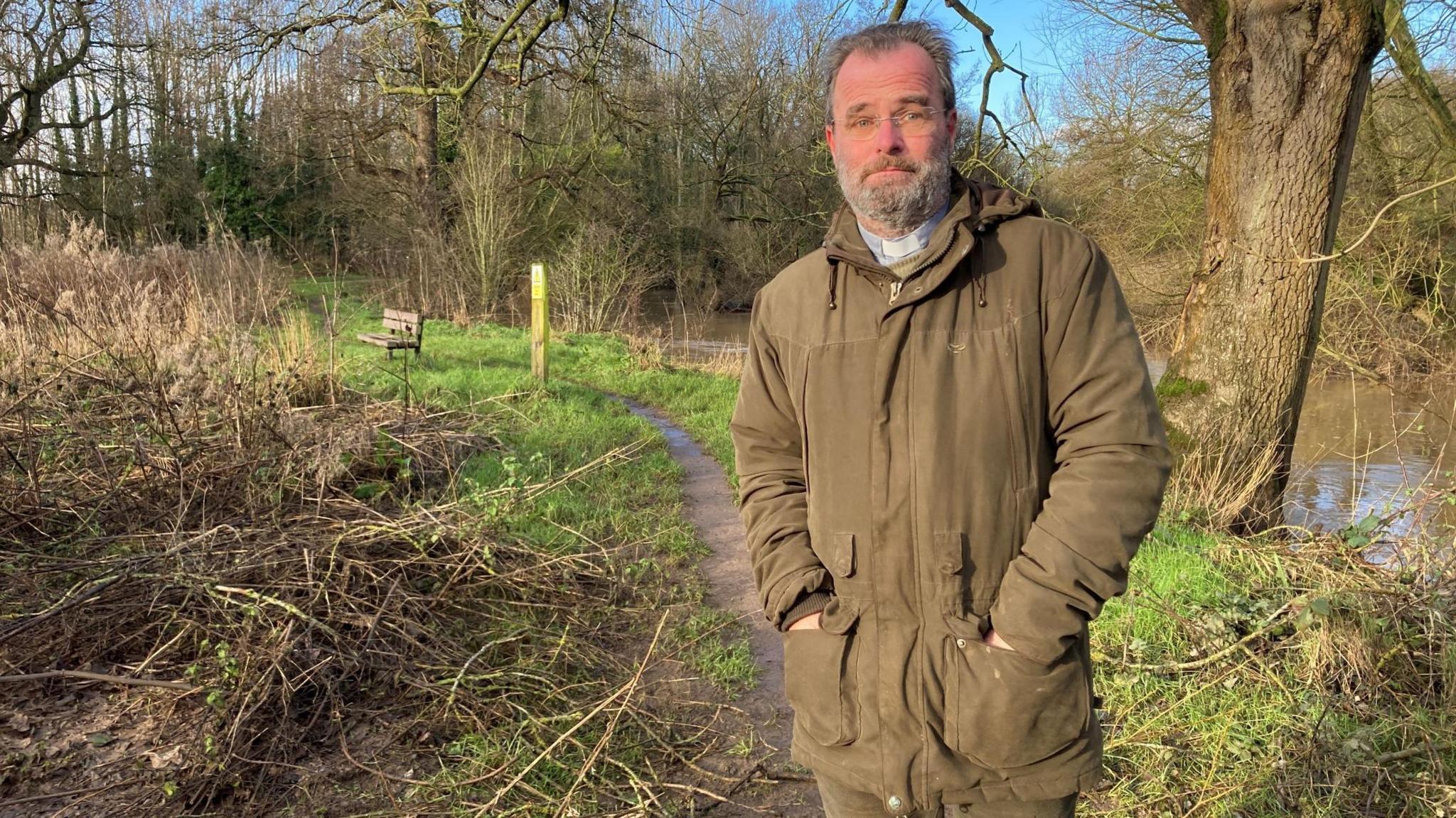 Vicar Paul Cawthorne is standing on a footpath with the River Roden on the right. On the left is flattened undergrowth, with traces of toilet paper.   