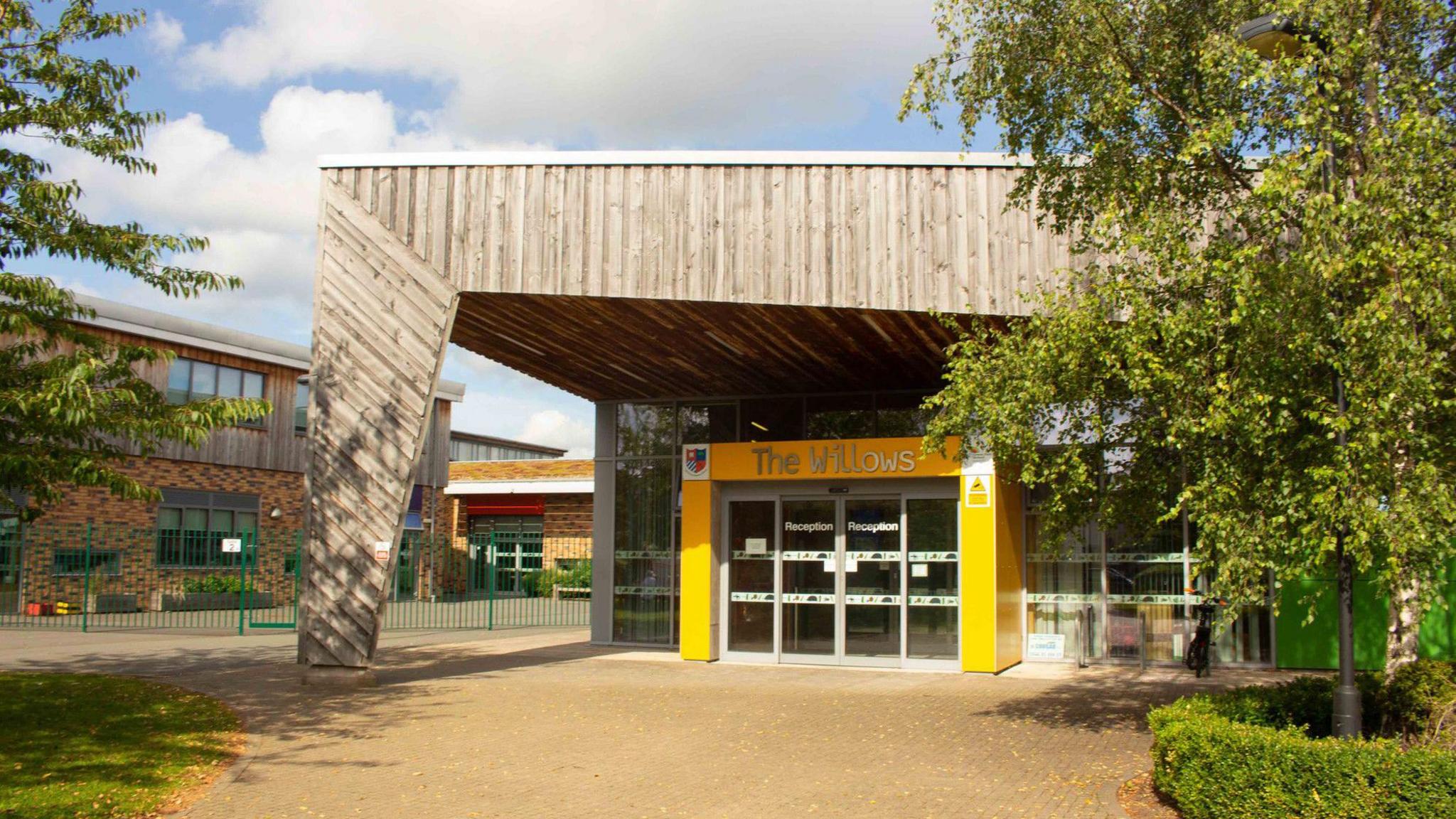 The entrance to a modern school building. Columns are painted yellow around a glass door, with a large wood clad pillar and roof. Willows is written in silver letters above the door, with reception written on the glass. Other school buildings can be seen to the left, with trees and bushes lining the path.