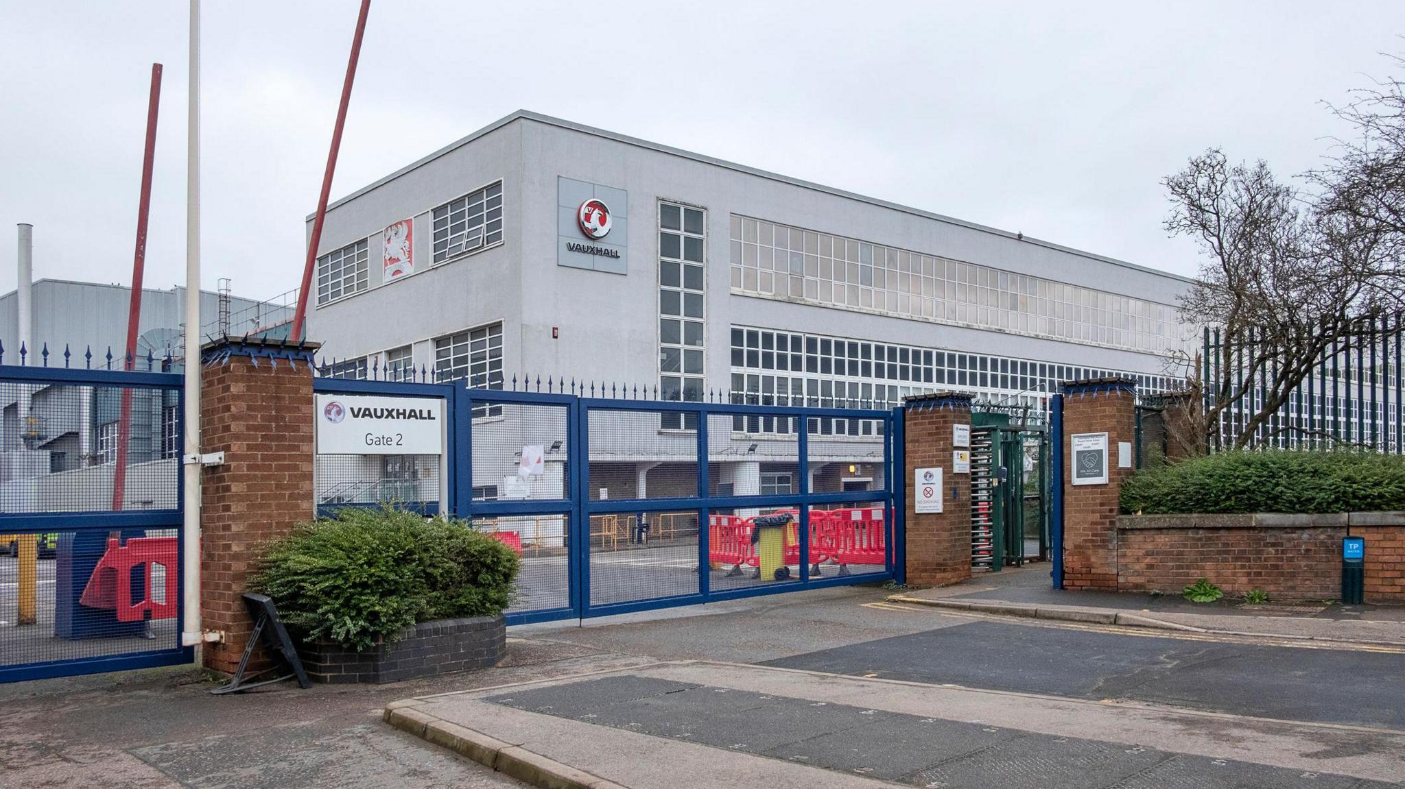 The entrance to the assembly plant of the automotive manufacturing company IBC Vehicles Ltd, operating under Vauxhall Luton and producing light commercial vehicles, in December 2021. It shows blue metal gates, red barriers, and a grey building with lots of windows 