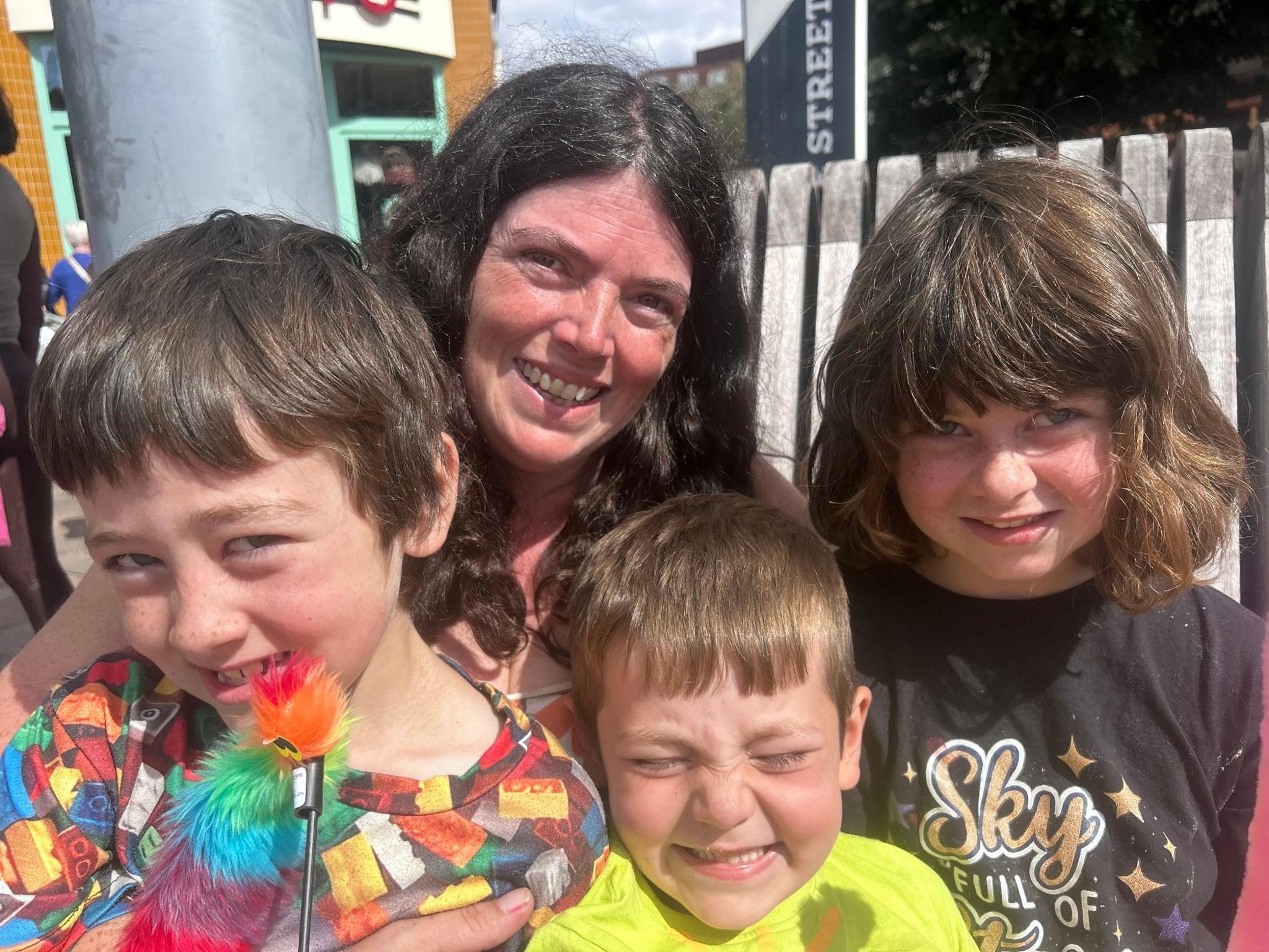 A woman smiles as she stands behind her three young children. She has long black hair. To the left, a boy wears a colourful Lego-themed shirt and has brown hair. In the centre, a younger boy has short fair hair and wears a yellow shirt. His face is scrunched into a smile. To the right, a girl has long brown hair and wears a black top with gold stars and lettering.