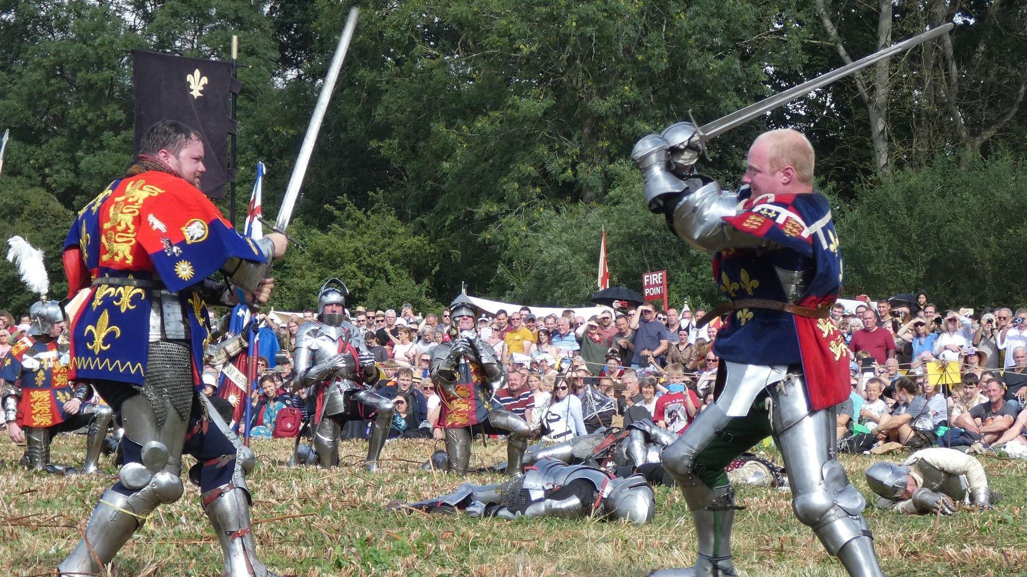 Two men in medieval armour fight with swords while some of playing dead on the ground and a crowd watches