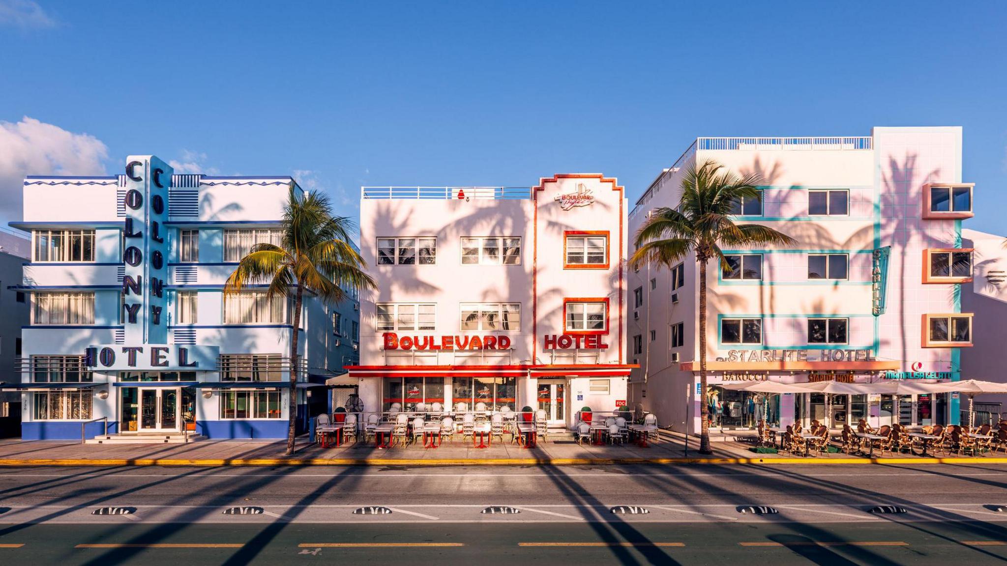 A row of brightly coloured art deco hotels pictured against a clear blue sky with palm trees in front