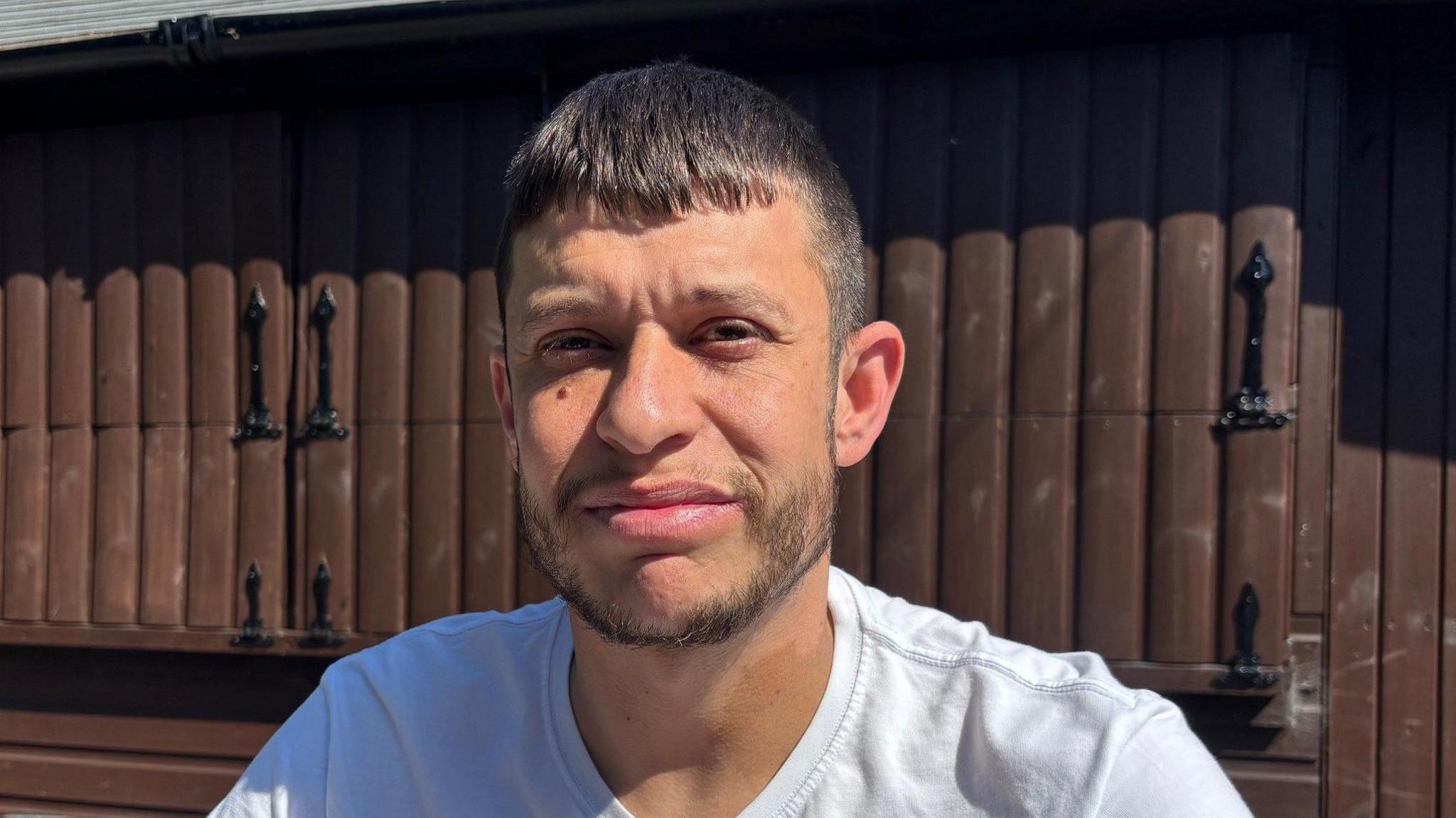 Arwel faces the camera, wearing a white t-shirt. He has short dark hair and stubble and is stood in front of a large wooden shed-type structure.
