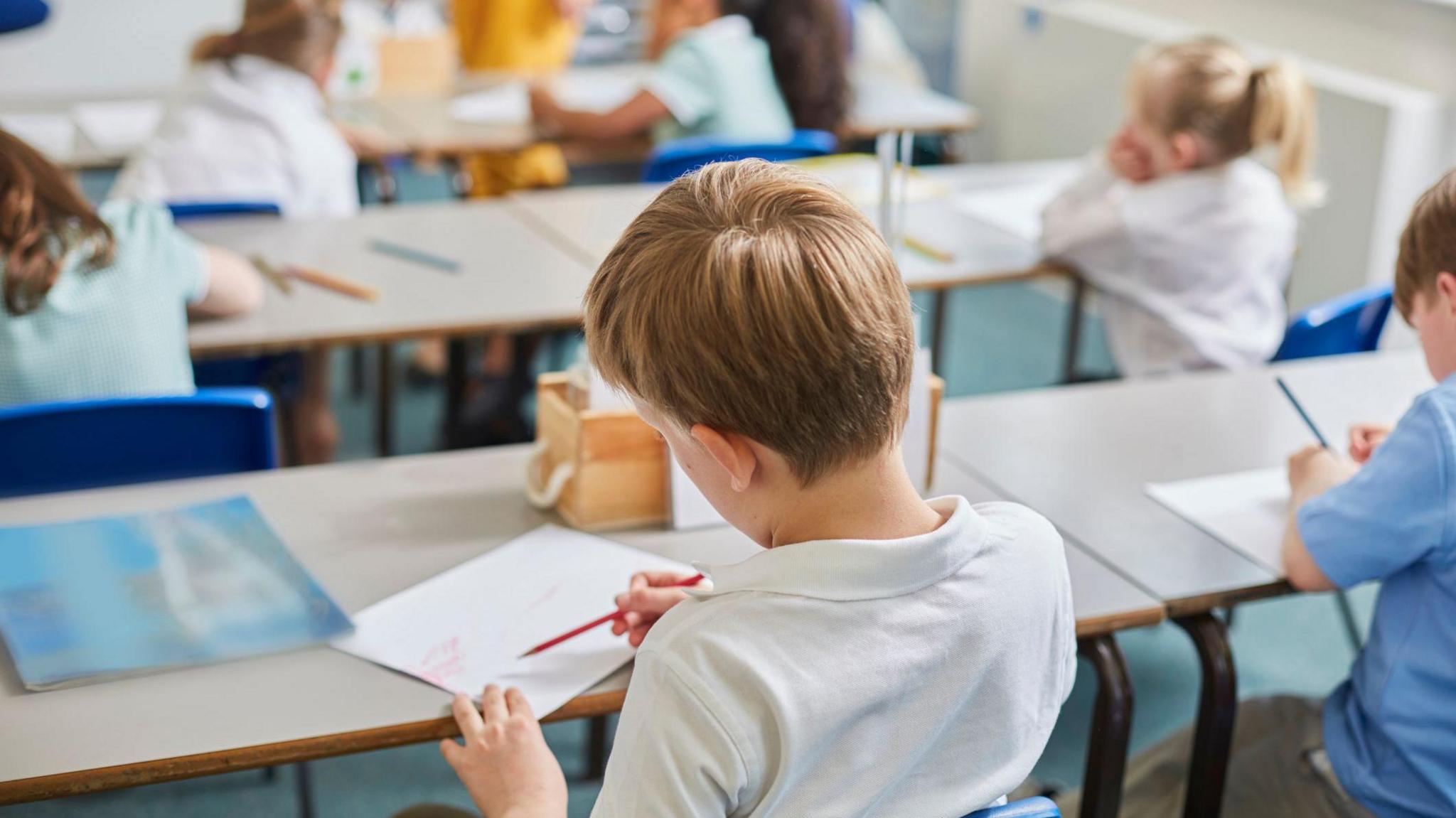 A boy is siting at a desk in a classroom. He has blonde hair and is drawing with a pencil on a piece of paper. Around him are other children at their desks.