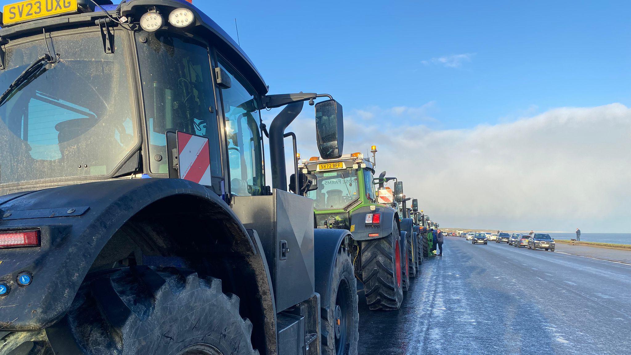 In the foreground of the image is the back of a tractor parked behind a green tractor. The sea is in the background. There's a number of parked cars on the other side of the road.