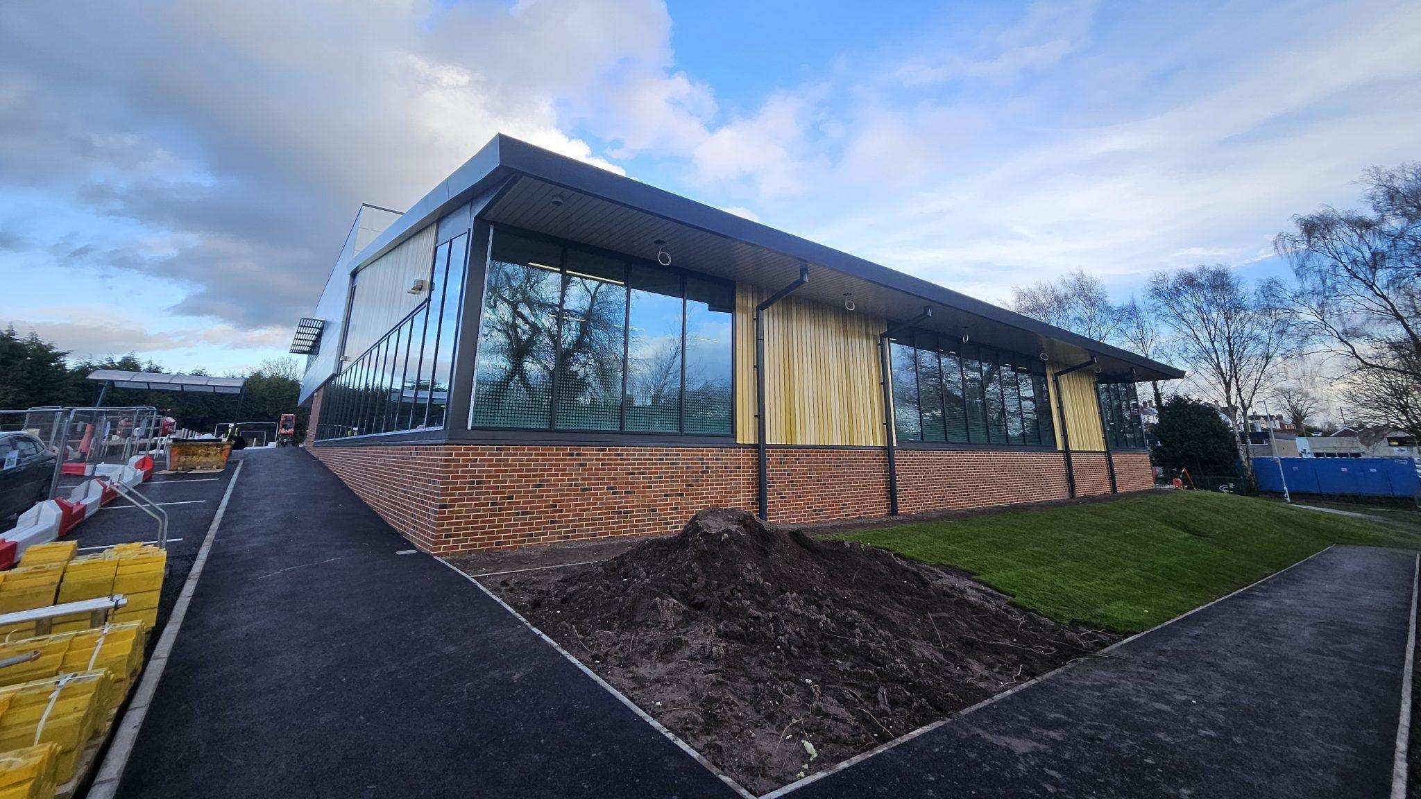 A rectangular building which has red brick at the bottom then dark grey windows and light wooden slats. The front of the building is not finished, there is a patch of grass on the right and a pile of soil on the left. The perimeter is still fenced off as construction work is ongoing. The sky is blue and cloudy in the background. 