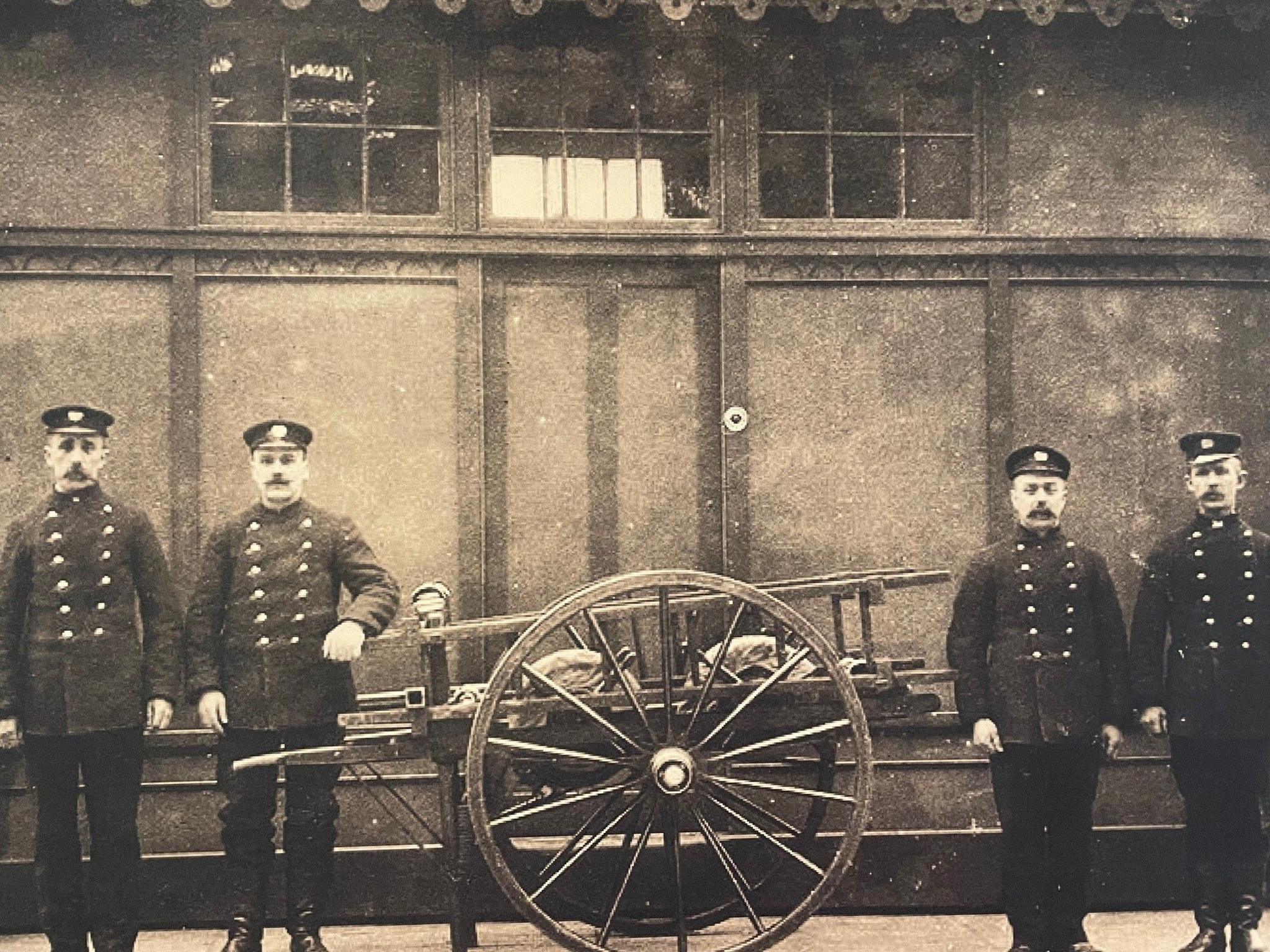 An archive shot of four firemen in uniform with a small wooden fire appliance contraption. The men are wearing tunic uniforms with lots of buttons and soft caps. They all have moustaches