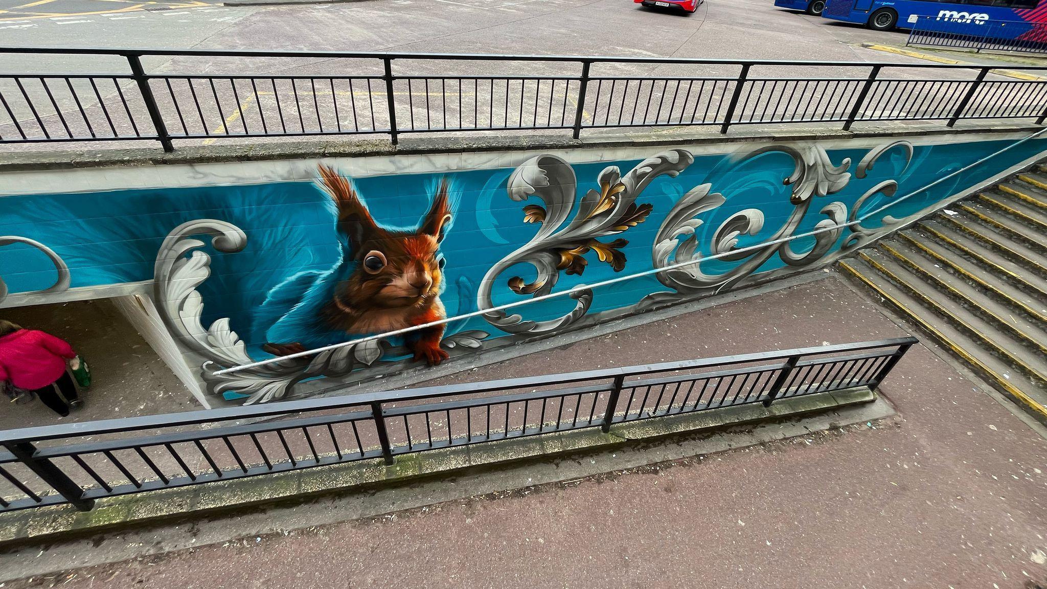 A blue mural with a red squirrel on the wall of the entrance to an underpass