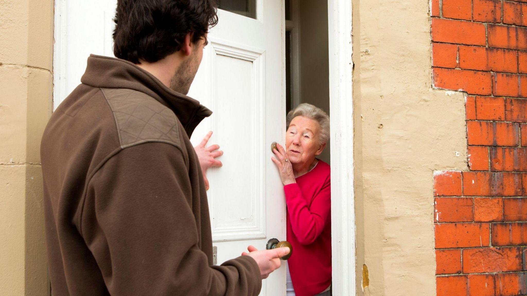 A man at the door, answered by an older woman. He is wearing a brown top and she is wearing a red top. The door is white, and there is brick around the frame. He has one hand on the door and she looks worried.