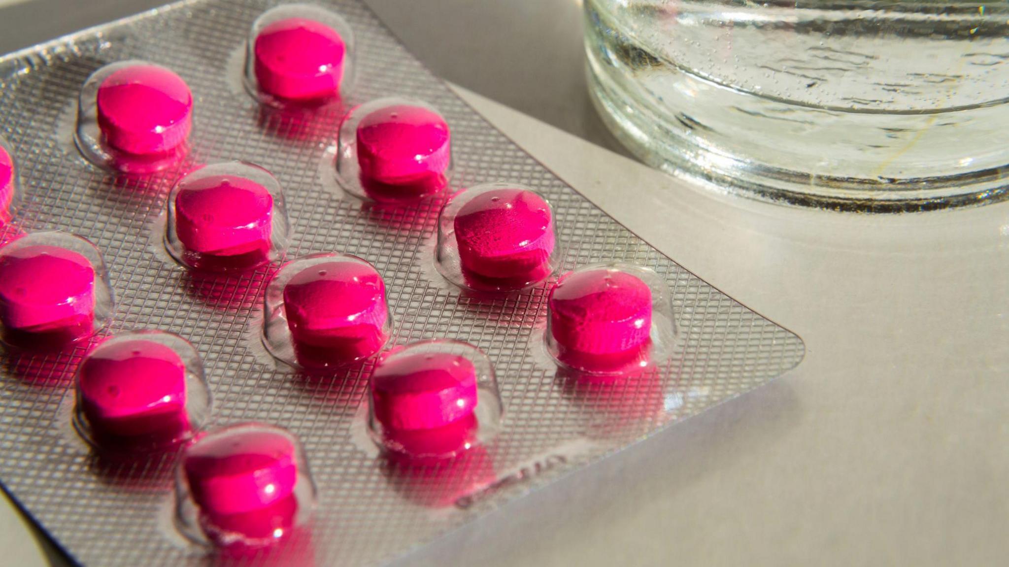 A packet of pink tablets, on a counter, by a glass of water. 
