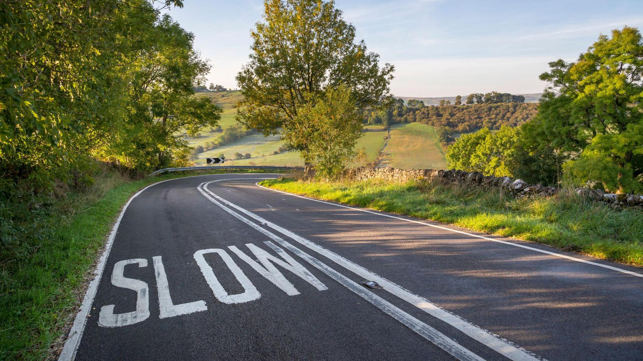 A country road at Crowdecote near Buxton in the English countryside. The road bends around to the right, with a slow sign visible. It is a beautiful bright day, with fields and trees visible. Very English looking countryside. 