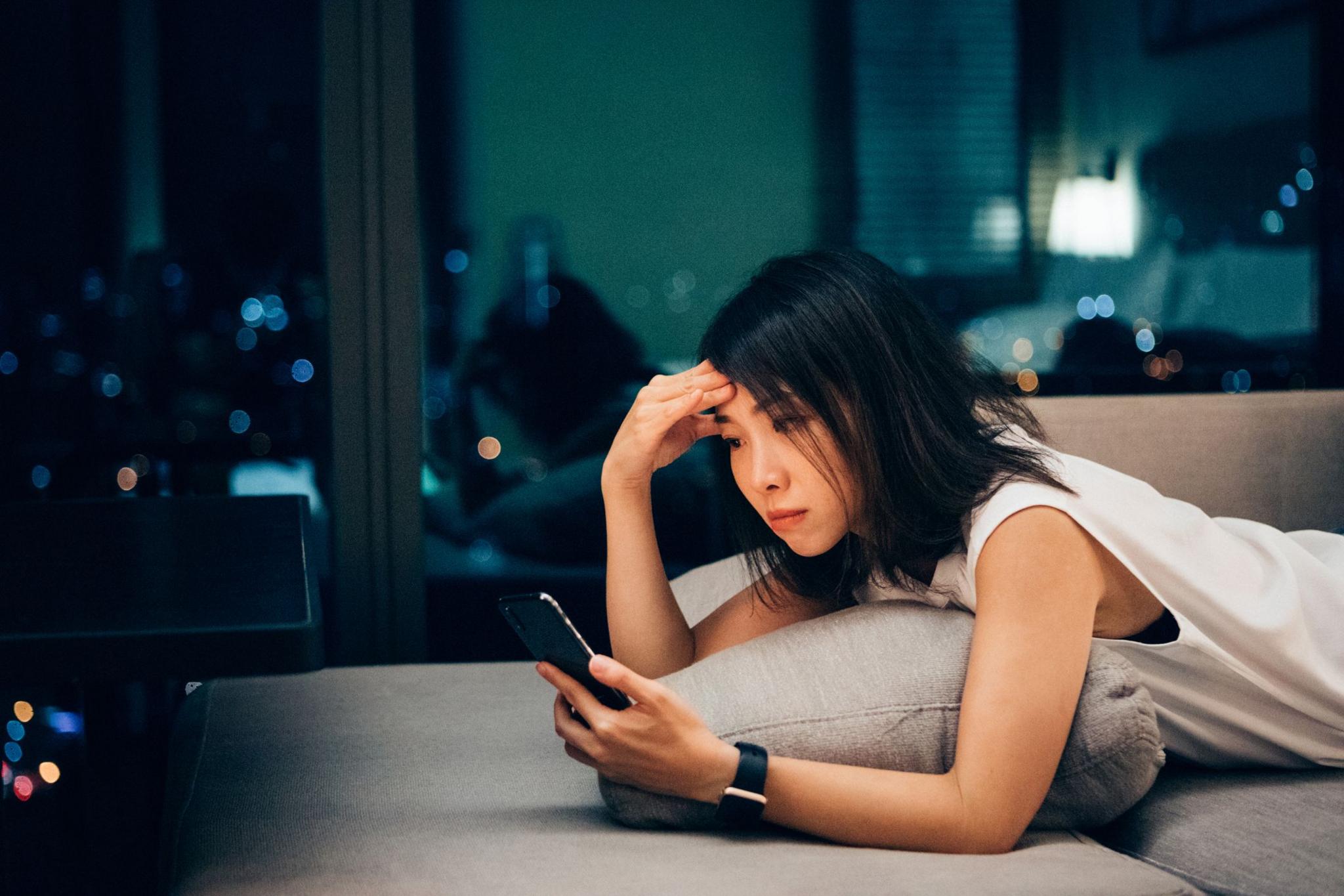 A woman lies on her front on a lounge, holding a phone in one hand and pressing her brow with the other