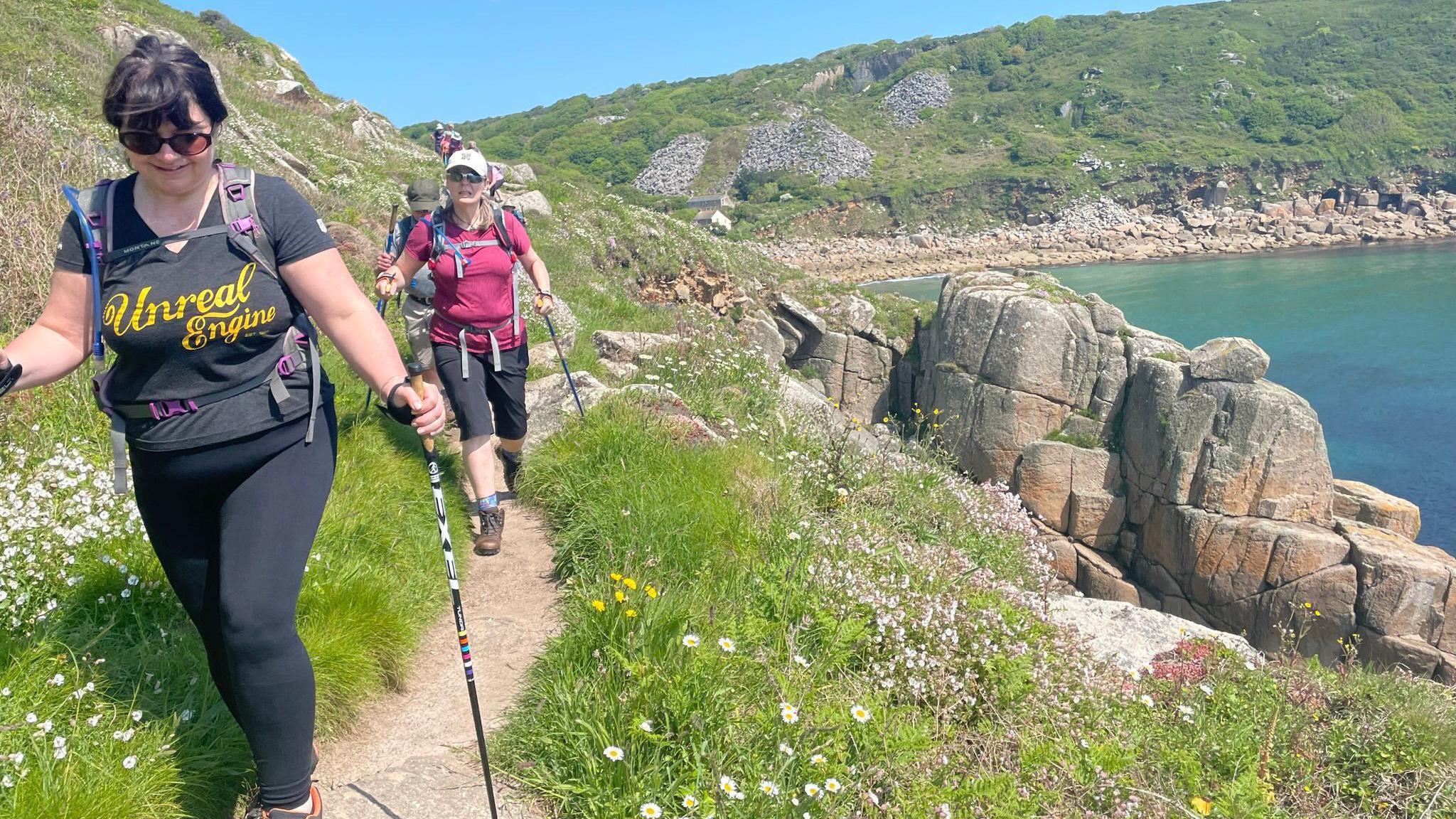 A group of Nordic walkers go along a coastal path in Lamorna so Land's End in Cornwall. The woman at the front of the group has a black T-shirt on with "unreal engine" written in yellow letters. The woman behind her has a burgundy top on and a white baseball cap. All the walkers have Nordic walking poles with them. The sea is on the right of the photo. It is a sunny day.