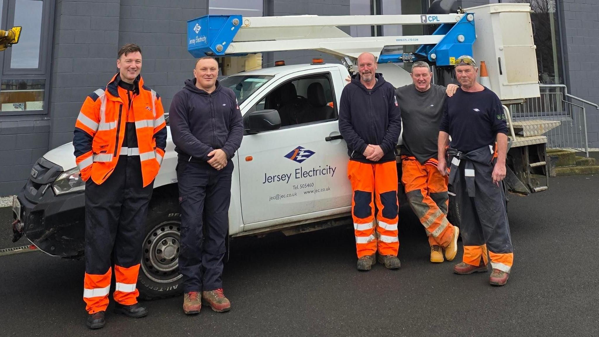 Jersey Electricity engineers are standing in front of a parked white van. There are two men on the left, and three on the right. They are wearing navy boiler suits and orange high-viz garments.
