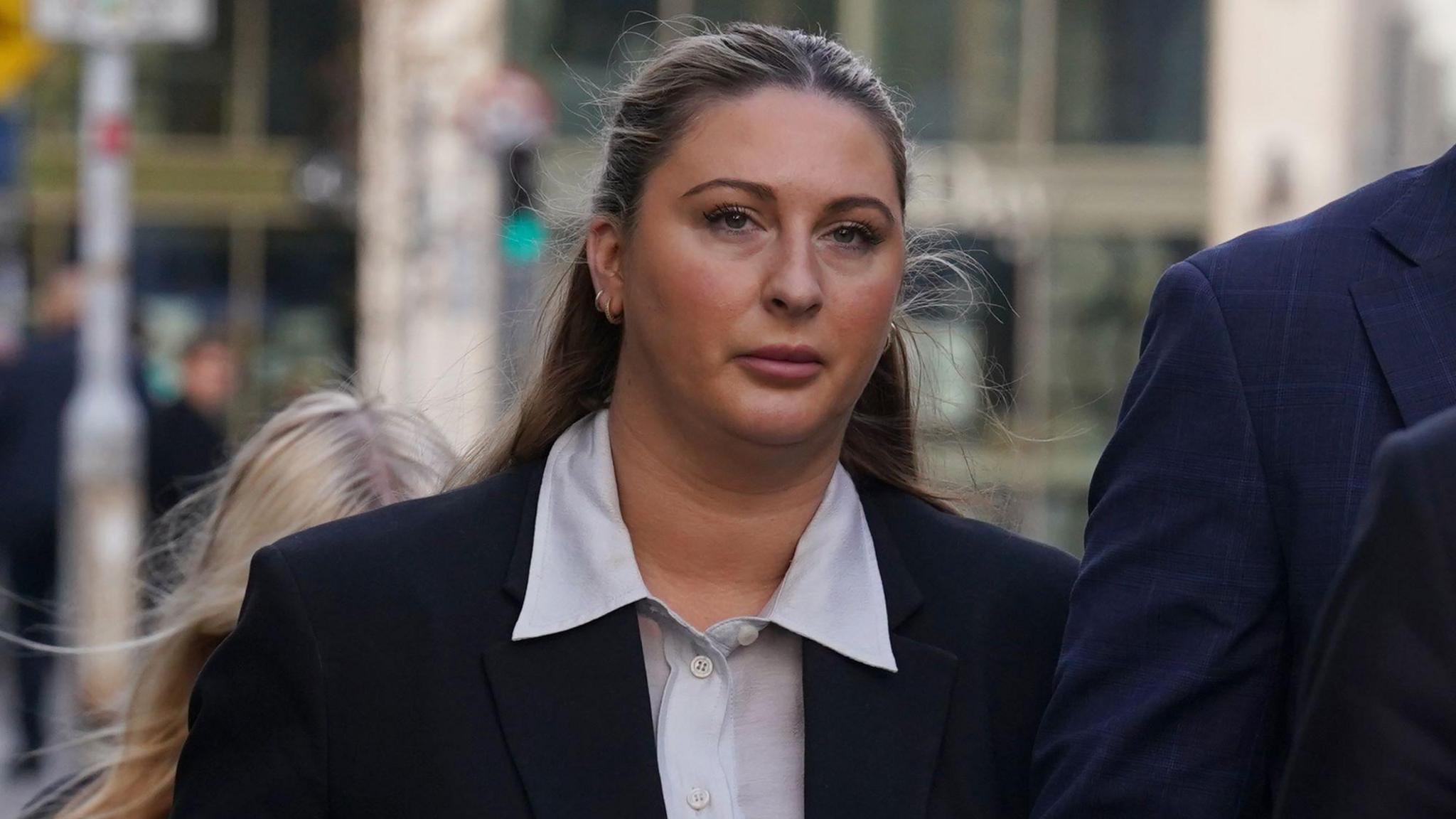 Ms Hand walking through the court gates, she has long blonde hair and is wearing makeup. She is wearing a black suit jacket with a blue shirt.