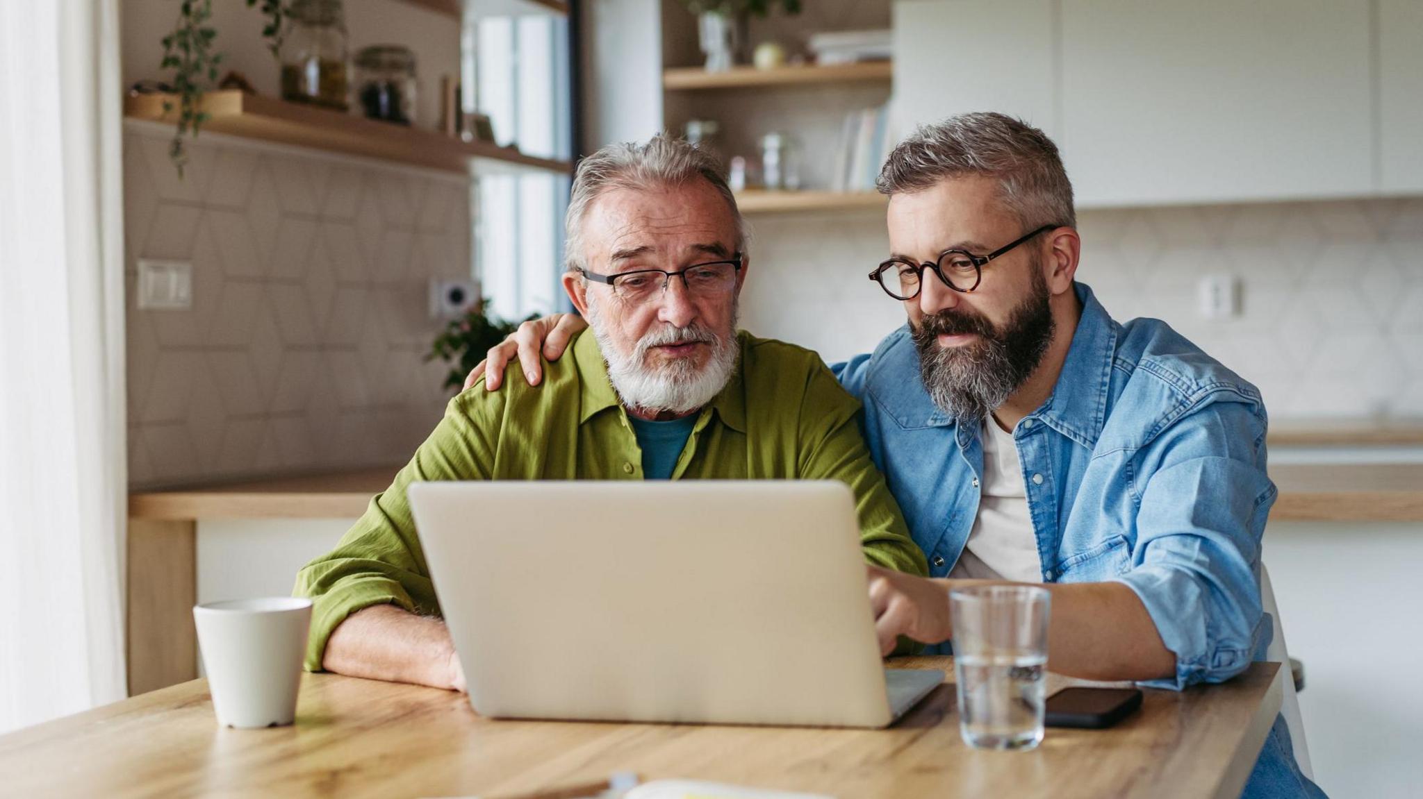 An older man with glasses wearing a green shirt next to a younger man with a blue shirt sat in front of a laptop.