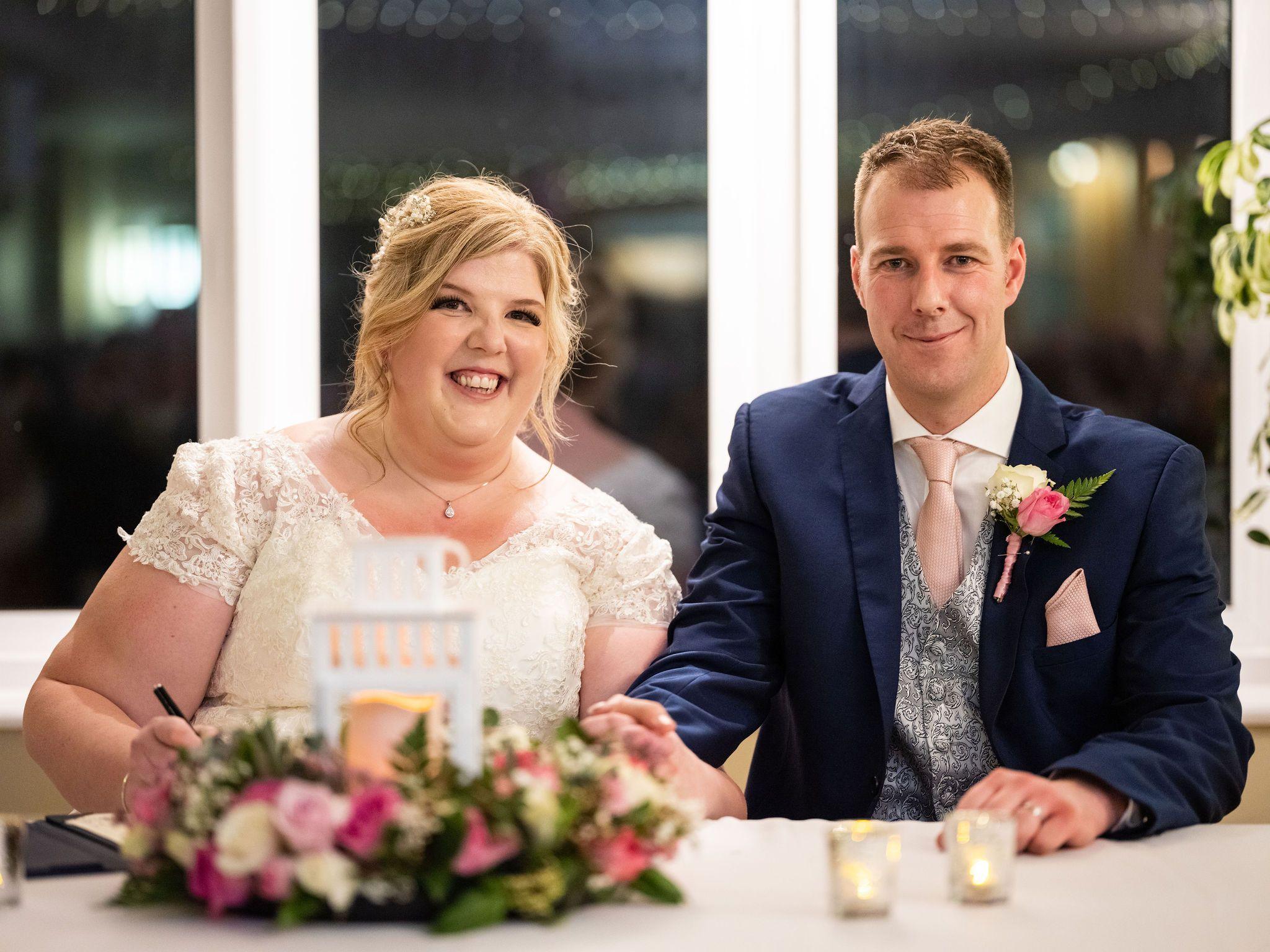 Amy Campbell is on the left wearing a lacy wedding dress holding a pen. Her husband is on the right wearing a navy suit, silk waistcoat and pink handkerchief and tie. There are flowers in the foreground. 