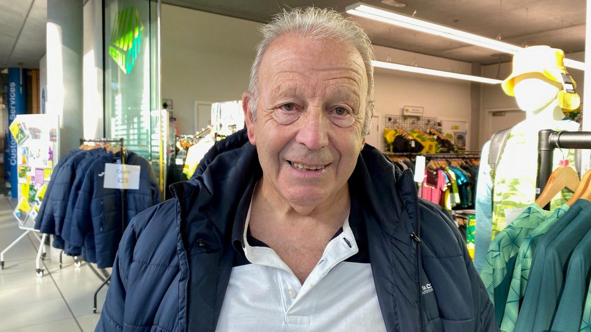 An older man wears a black padded jacket and white casual shirt. Behind him are clothing rails.