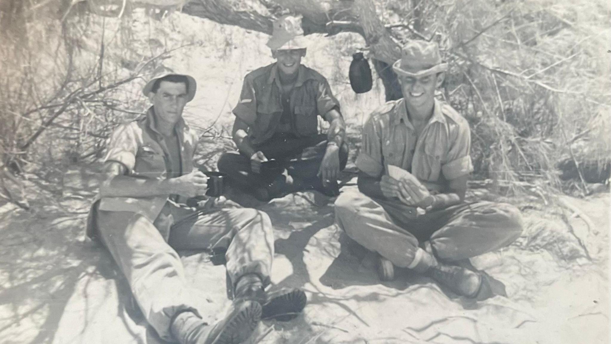 Black and white photo of three men in khaki military uniform with hats, smiling at camera, sitting on rough dusty ground beneath dry brush.