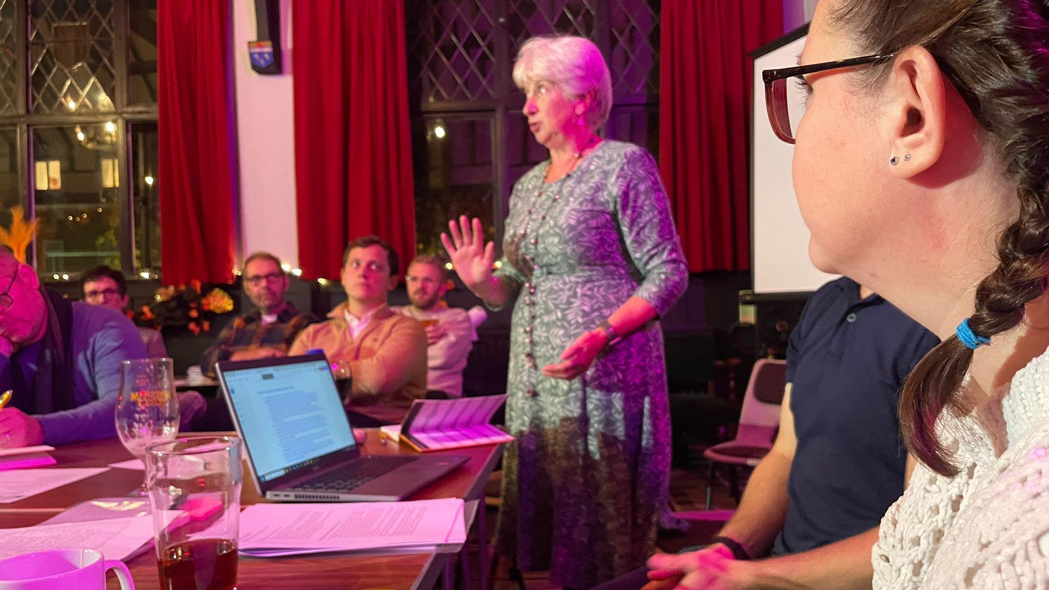 Menyw yn sefyll ac yn siarad i gynulleidfa o gwmpas bwrdd, gyda chluniadur a phapur o'i blaen. Mae ganddi wallt gwyn ac mae hi'n gwisgo ffrog werdd, ac mae'r cynulleidfa'n edrych ac yn gwrando arni wrth iddi siarad.