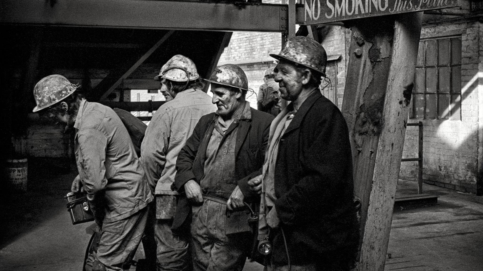 A black and white image of a group of miners at the end of their shift. They are all wearing overalls and hard hats and are covered in coal dust.