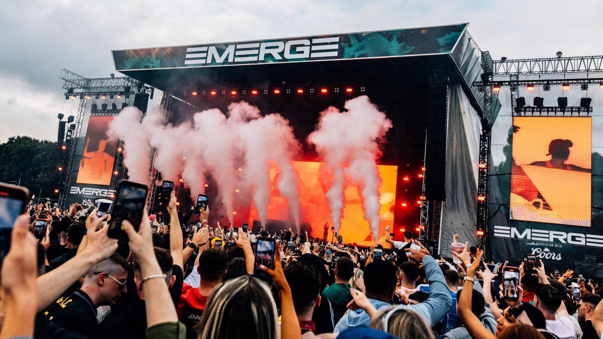 The main stage at Emerge. The stage has pink smoke shooting up from it with a large sign reading Emerge in capital letters. There is a big crowd of people and lots of them are videoing on their phones.