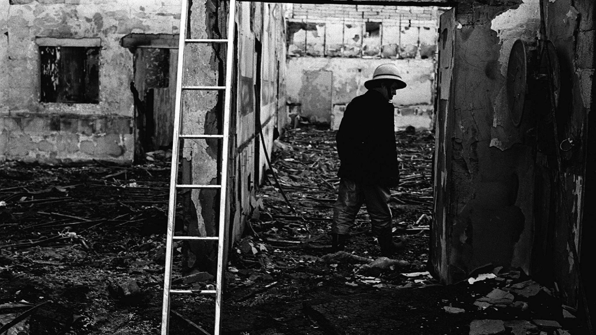 Black and white image of the bombed La Mon hotel, the brick building is covered in black with parts of walls fallen down, debris lies on the floor and a man stands by a ladder in the debris, he is wearing a dark jacket and trousers with wellington boots and a hard hat. 