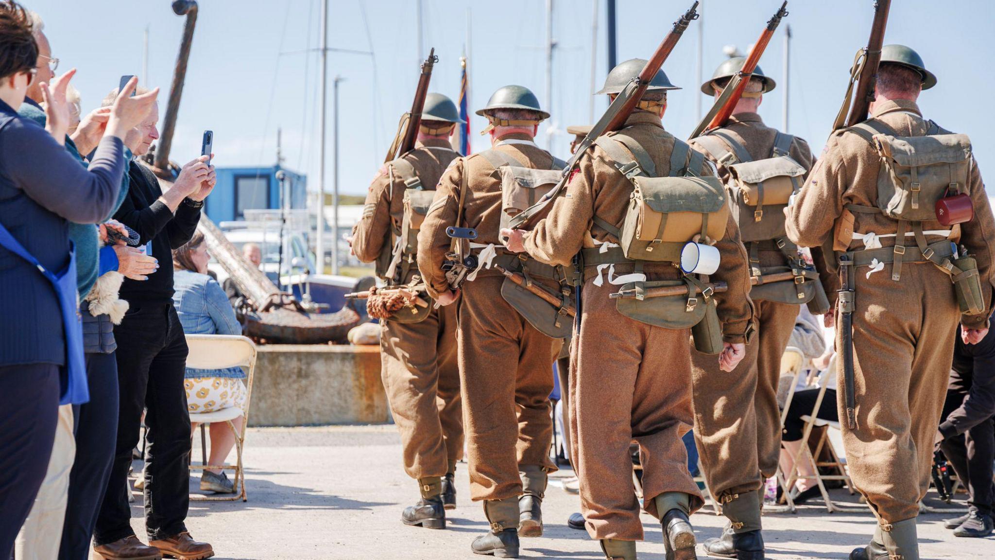 A group of soldiers in historic military uniform begin to march from the harbour while onlookers applaud. 