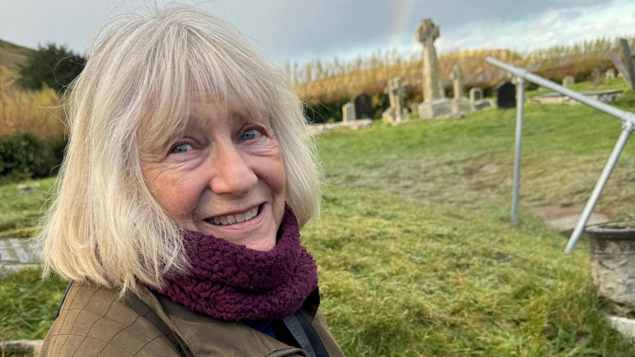 A woman with grey hair, a purple scarf and a brown coat standing in a grassy field with headstones in it