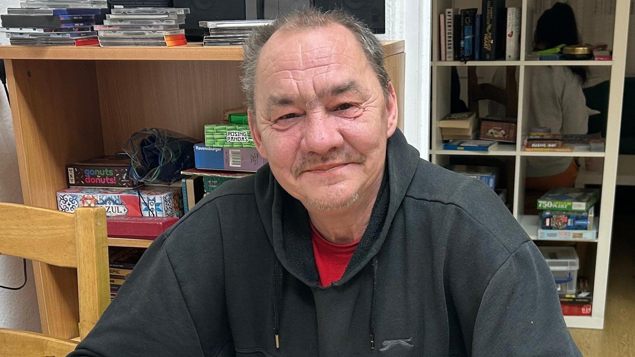 Slowomir, sitting in a room, looking at the camera, smiling, he is wearing a black top, with a red T-shirt underneath. There are shelves behind him with items on them. He has short hair, that is slightly greying. 