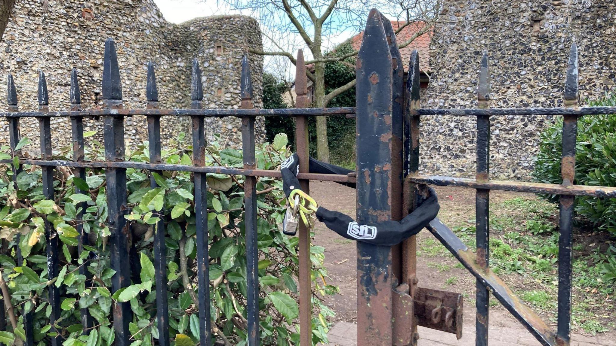 A black chain and padlock wrapped around a black metal fence. The fence is surrounded by bushes and behind the gate is a clearing flanked by two old stone walls.