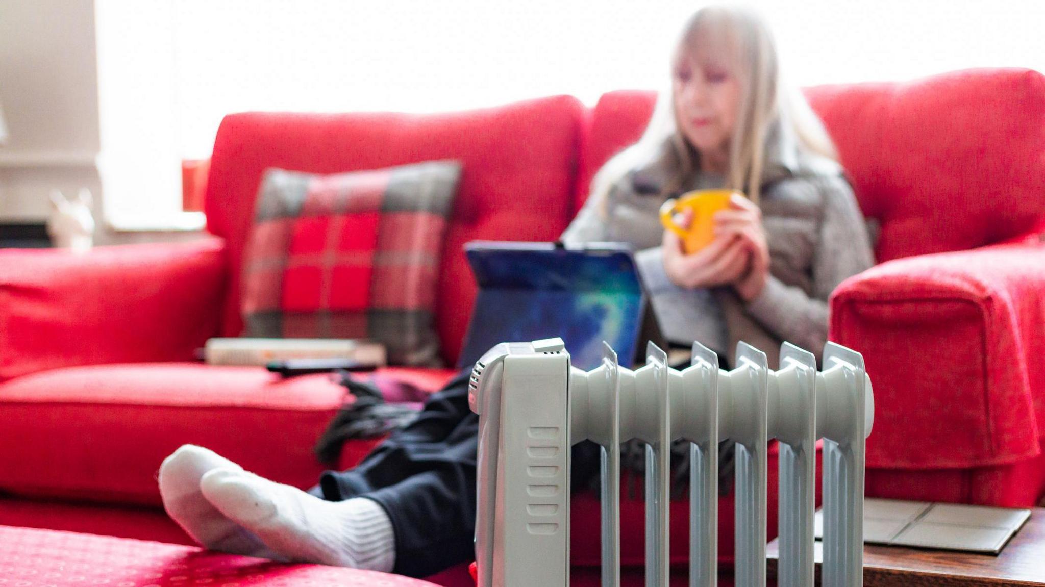 An older woman sits on a couch with a cup between her hands. She has her feet on a stool next to a heater