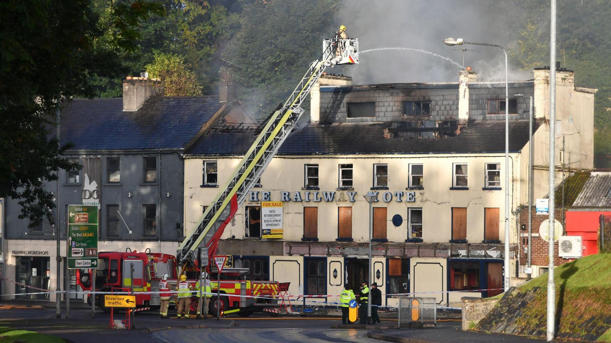 Scene of fire from a distance showing building with fire damage to the roof. A fire appliance uses a ladder to spread water over the fire.