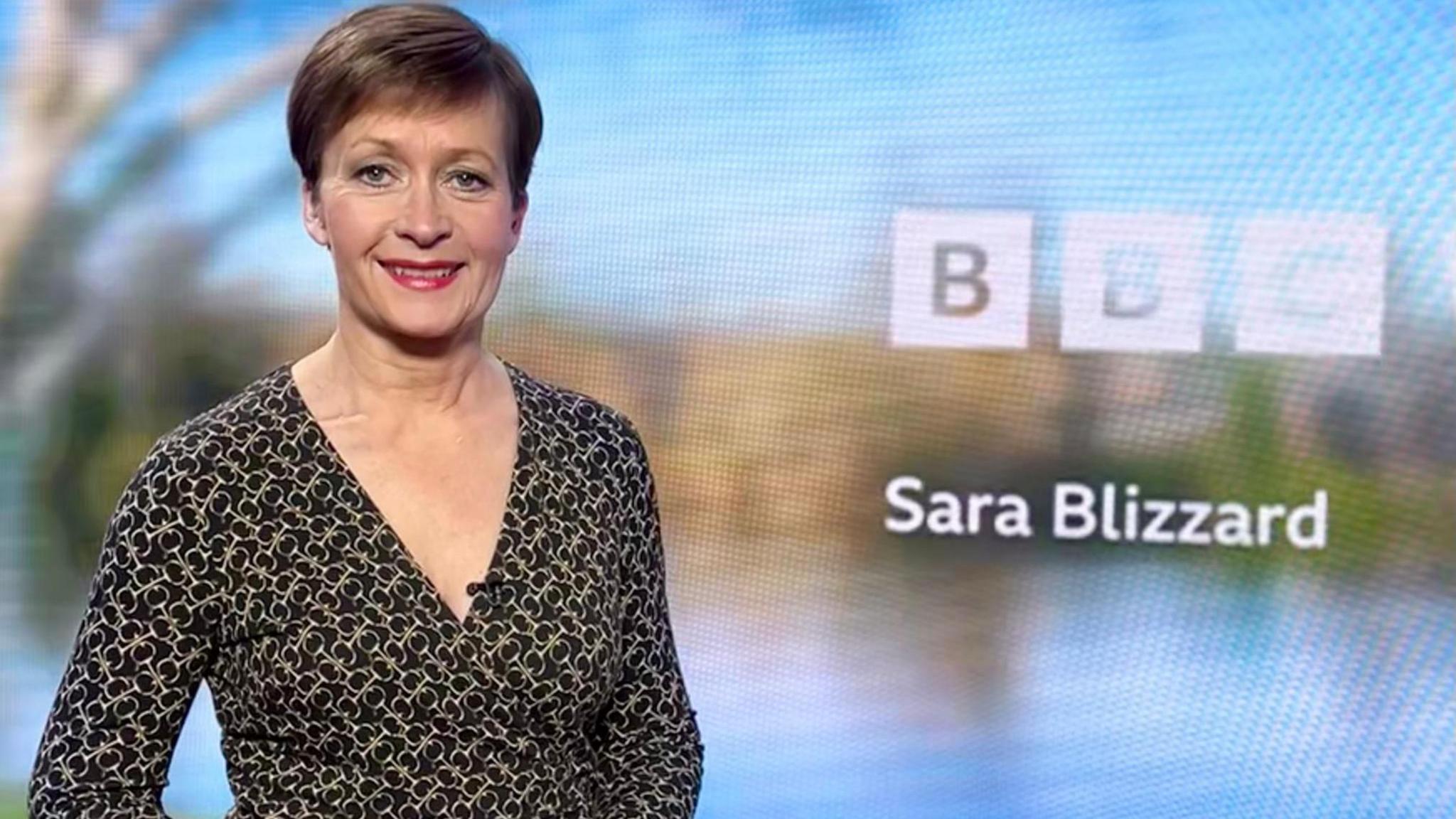 Sara Blizzard wearing a black patterned dress stood in front of a digital screen.