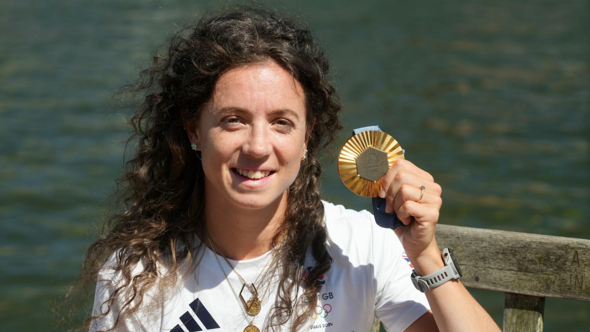 Imogen Grant smiling at the camera with long, dark curly hair. She is holding up her gold medal. 