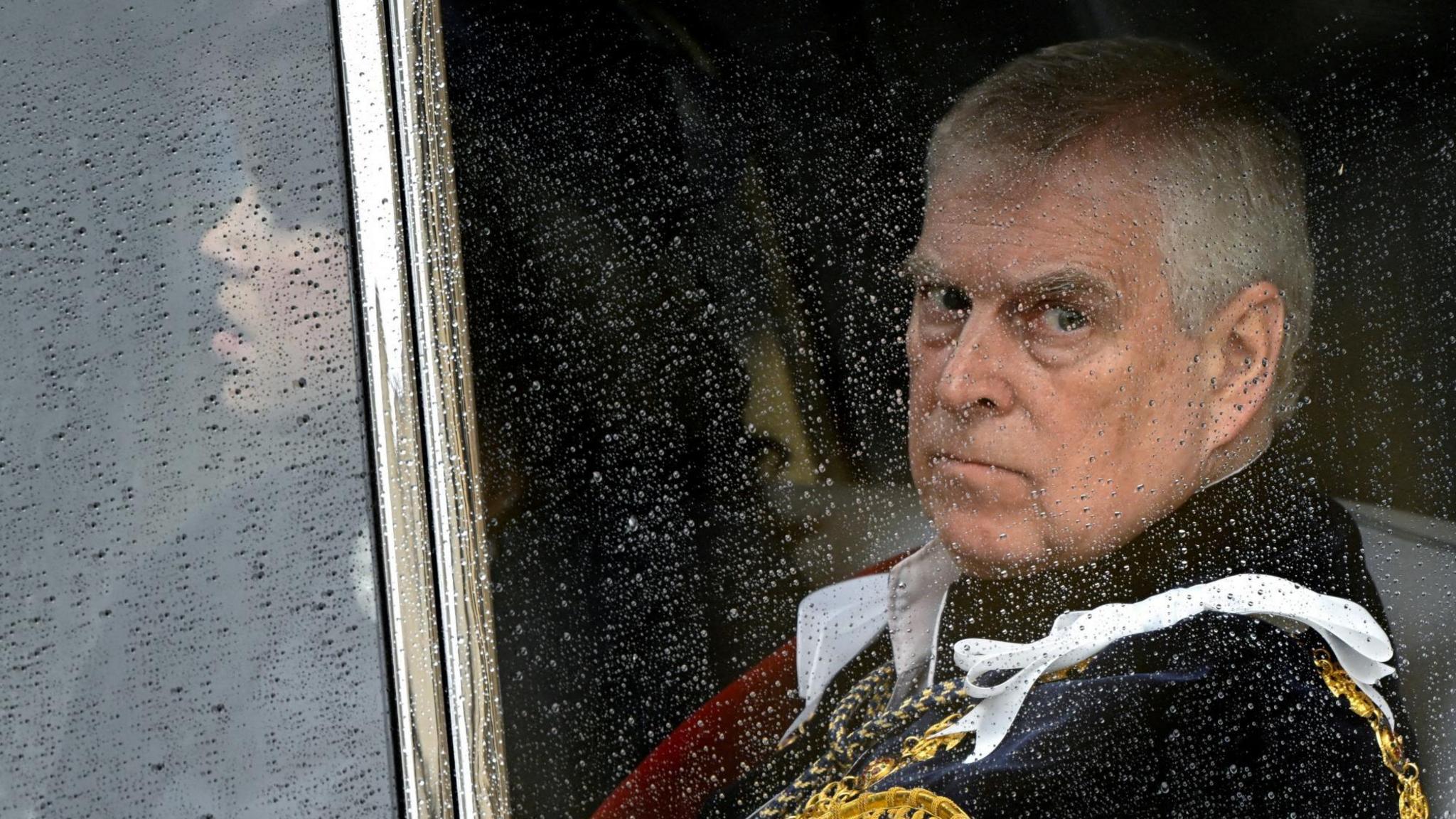 Prince Andrew, in royal robes, is pictured through a carriage window smattered with rain.