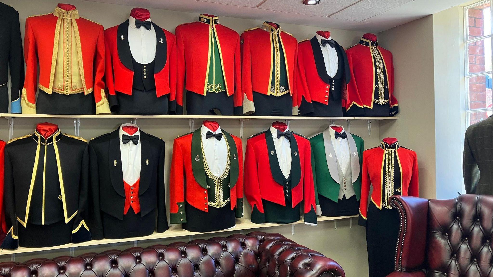 The tops halves of dress uniforms are seen on two shelves on a wall. The jackets are mostly red, some are black, and they have varying gold trims and some with black or green cloth underneath, some with high collars and others with bow ties at the collar.