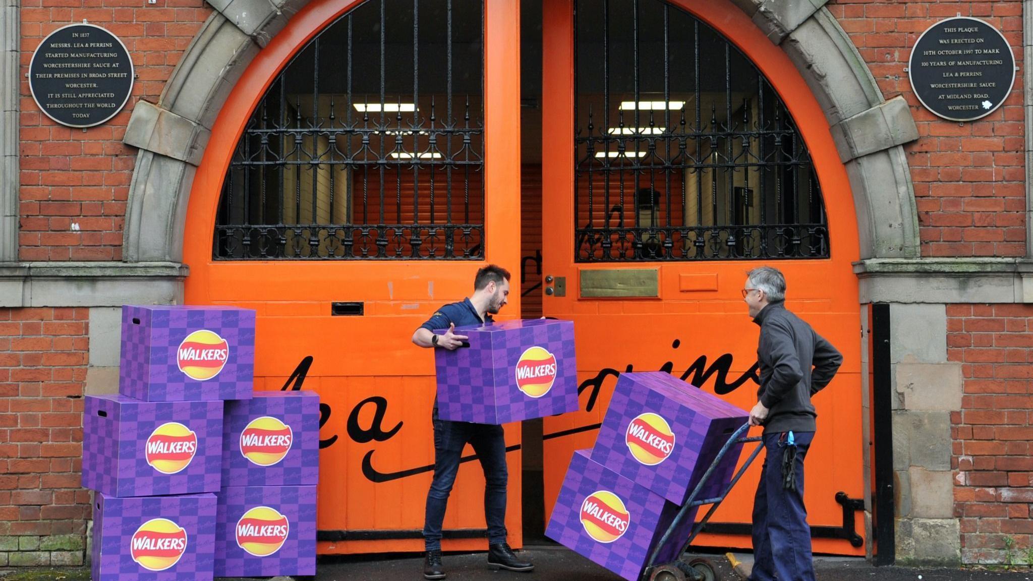 Purple boxes of Worcester sauce Walkers crisps being taken into the large orange doors of the Lea & Perrins site. One man is carrying a box and another has two boxes on a trolley. Black plaques with gold lettering flank each side of the door.