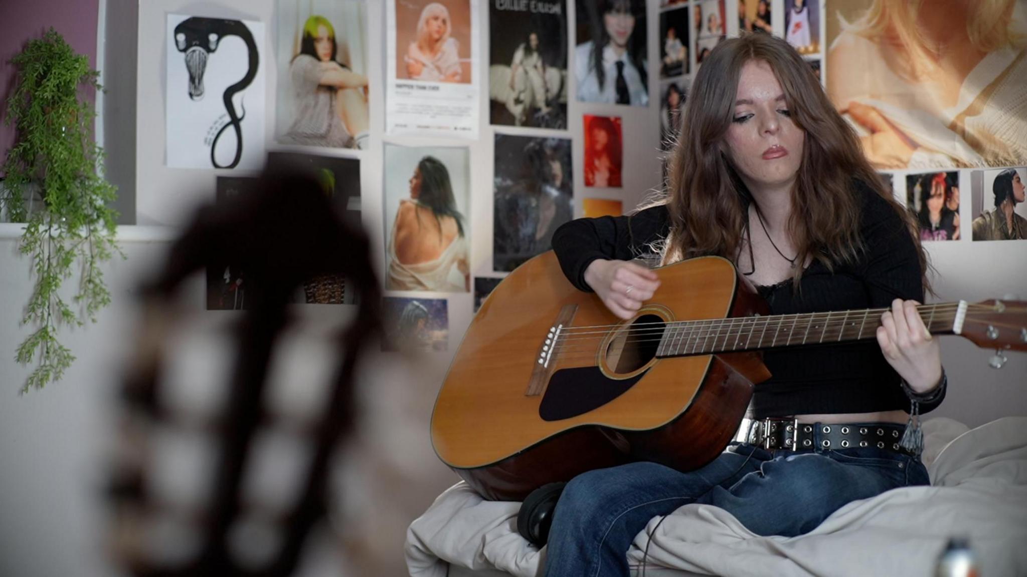 Amy sits on her bed in front of a range of Billie Eilish posters on her bedroom wall. She is holding a wooden acoustic guitar and is looking at her fingers playing it. There is a green plant to the left of the photo and another guitar to the left foreground. To the right Amy sits. She is wearing blue jeans, a black and silver-coloured belt and a black top.