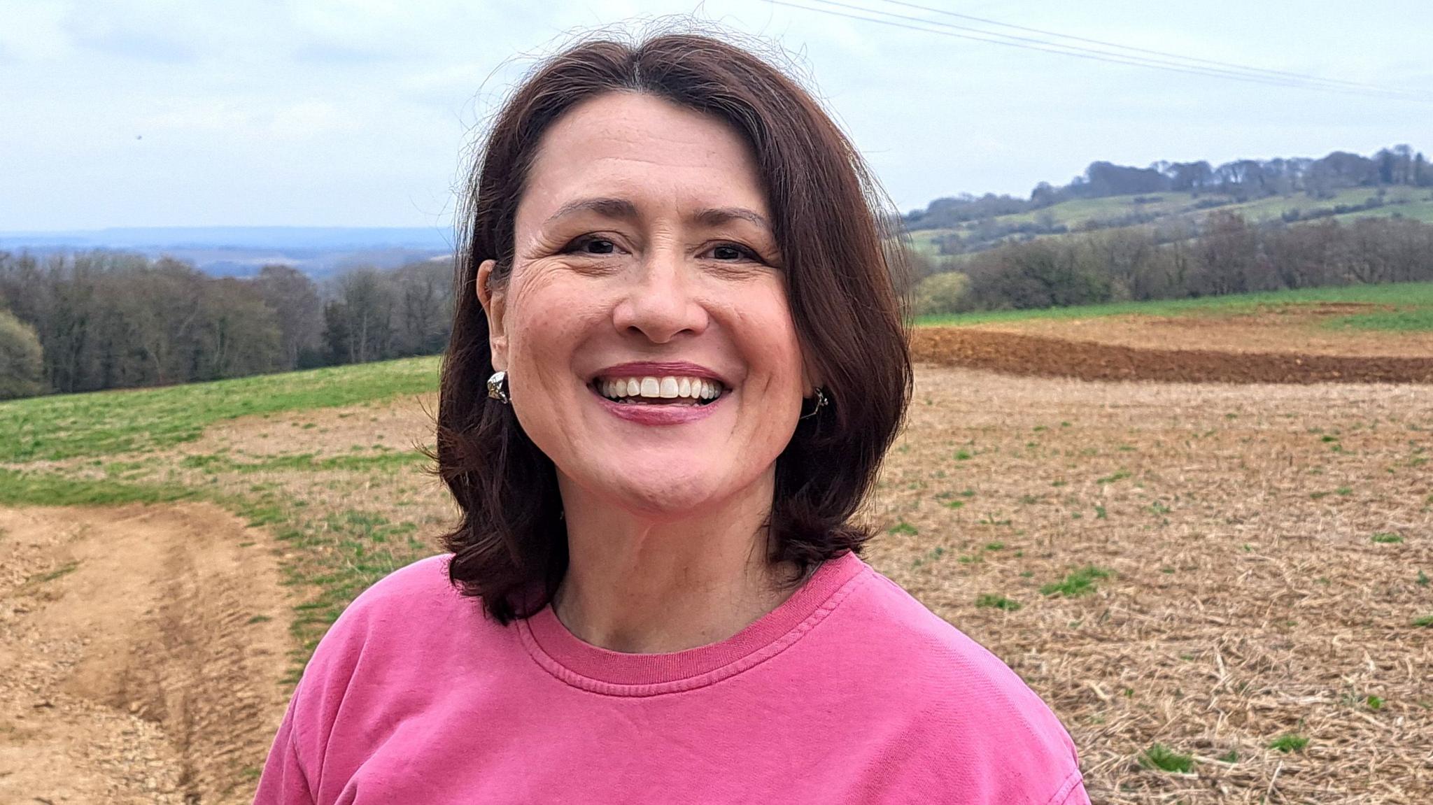 Georgia Elliott-Smith is smiling broadly in a field, with hills and cloudy skies behind her. She is wearing a pink jumper and has brown shoulder-length hair. 