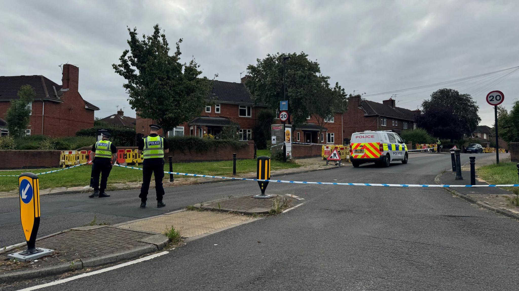 Two police officers, with their backs to the camera, guard a police cordon blocking off a residential street. A police van is within the cordon. 