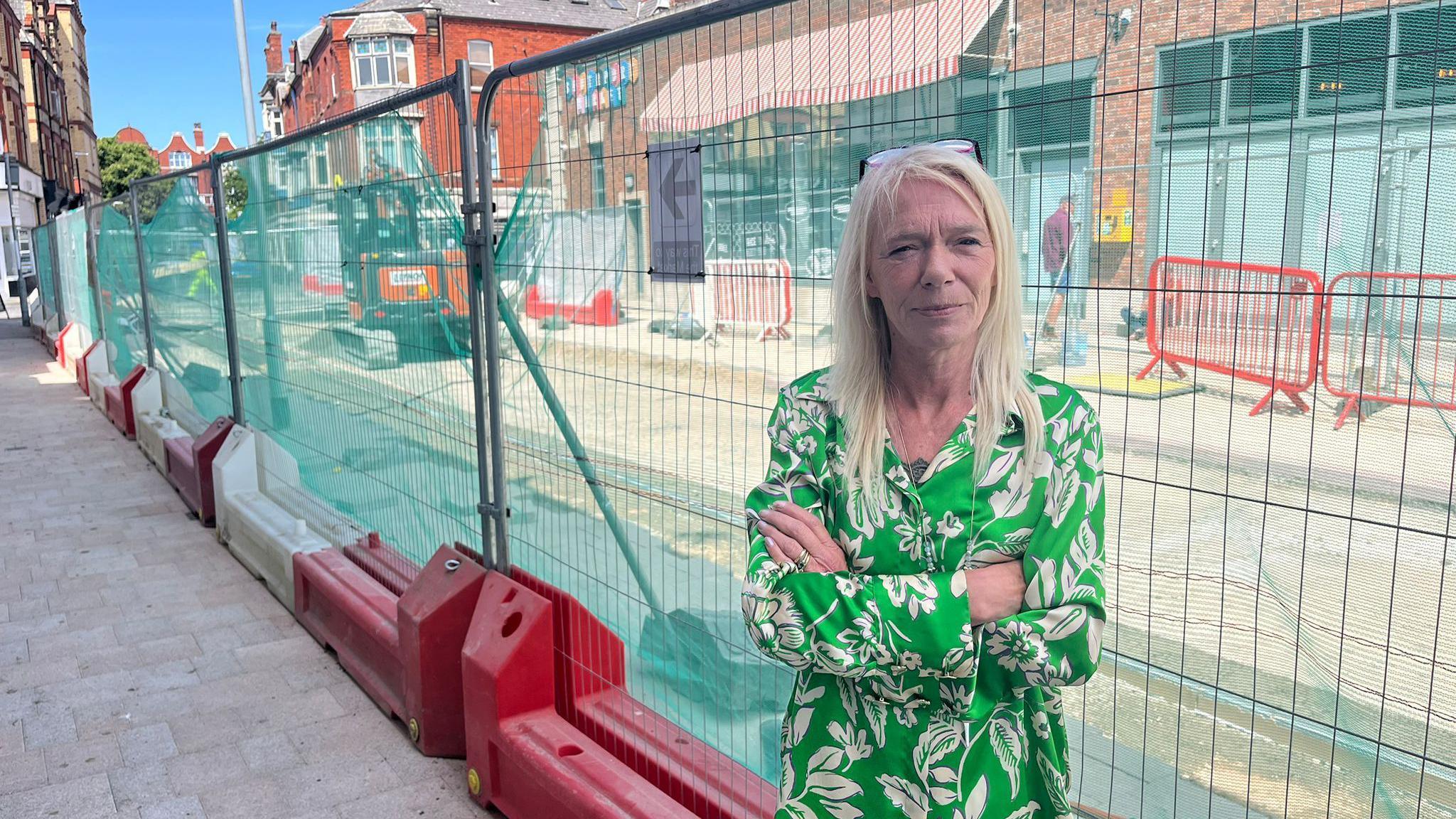 Tracy Jones is stood in the street next to large metal fencing with roadworks behind. She has long hair and is wearing a green and white floral shirt and has her arms folded.