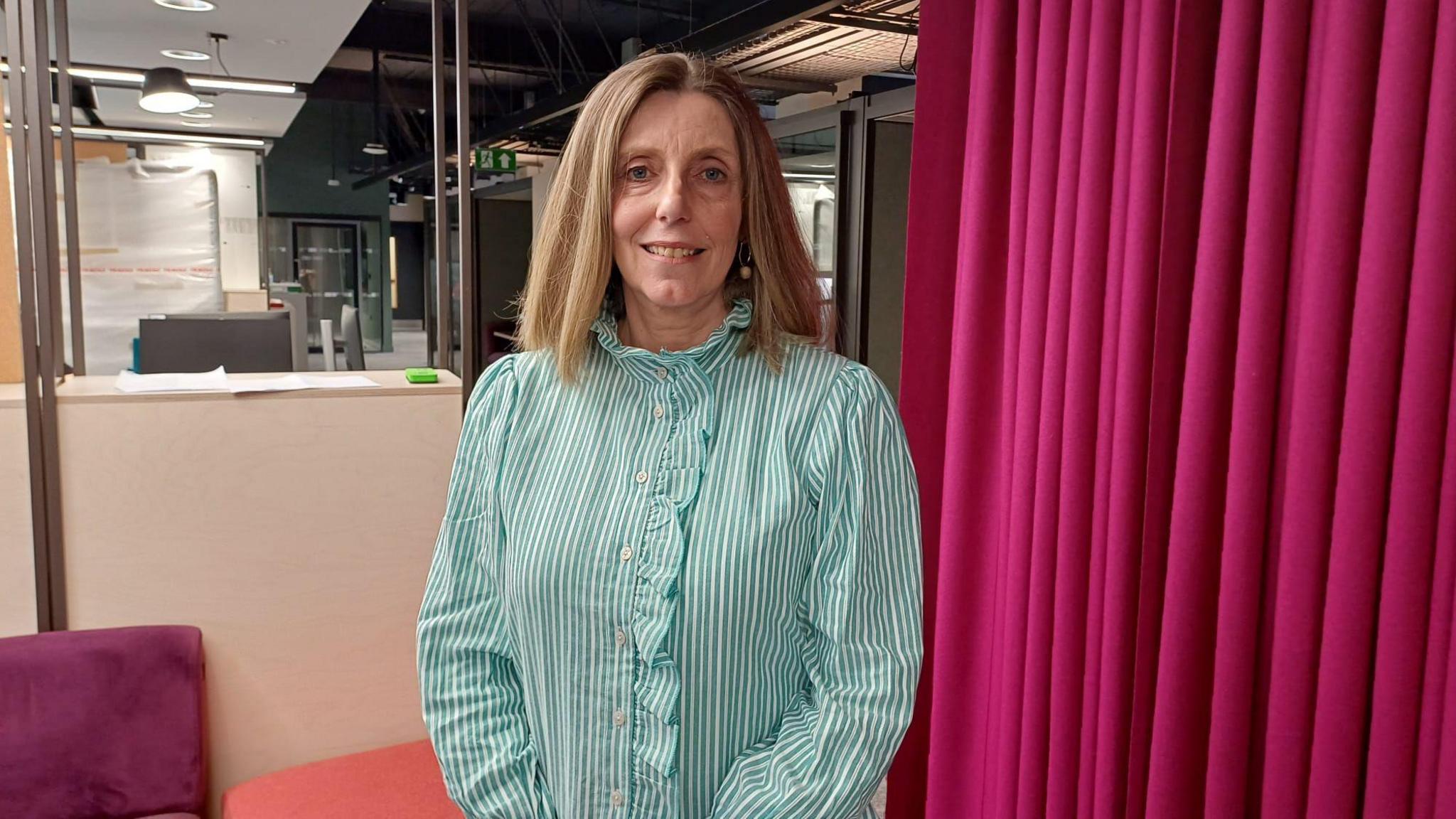 Gwyneth Jones is standing in an office. She is wearing a green and white stripped top and hoop earrings with a pendant on them. There is a pink curtain behind her.
