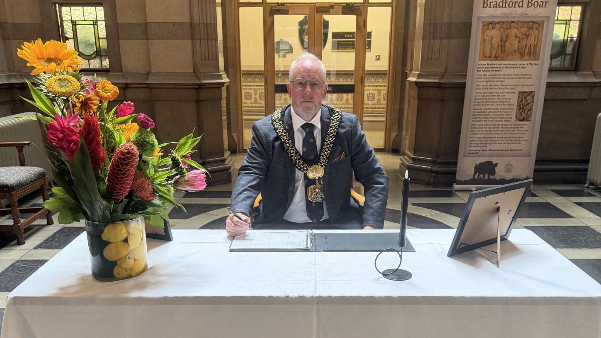 A man with short white hair wearing a suit and mayoral chains sits behind a white table with a large floral tribute on the left of the table. He is signing a book in front of him