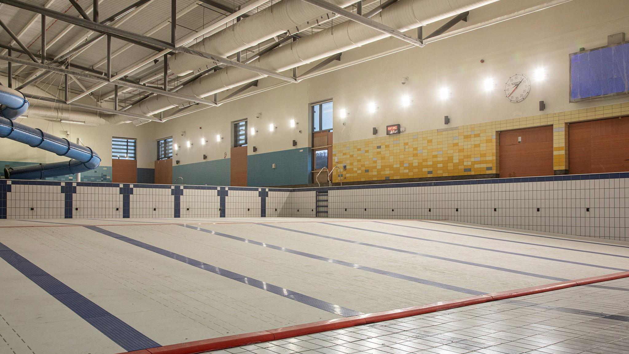 The empty main pool in Newry Leisure Centre photographed in 2024.  The pool has white tiles interspaced with blue tiles marking separate swimming lanes. A ceiling has visible pipe work and a blue flume is seen at the left of the photo. 