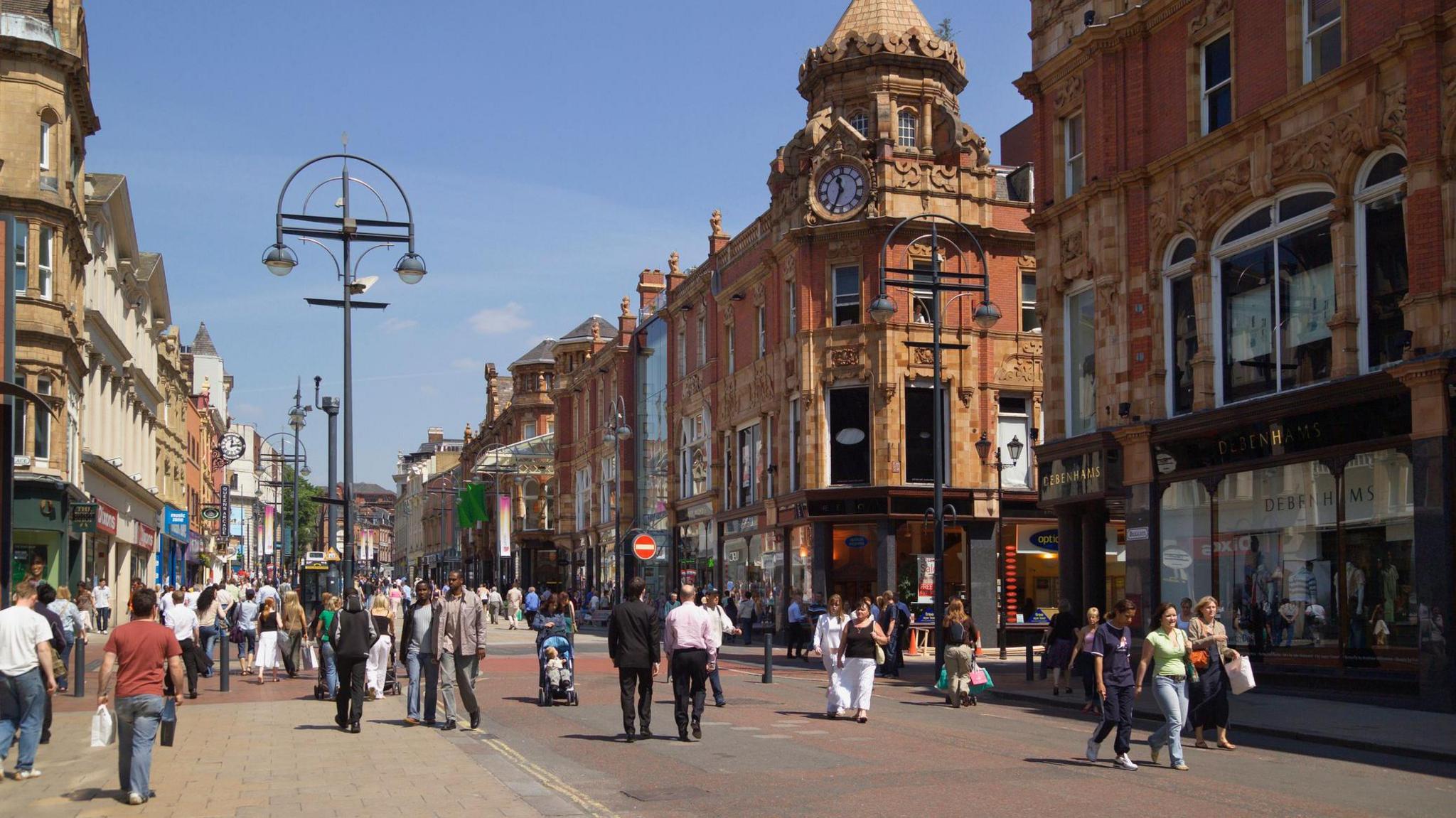 A bustling pedestrian street in a UK city centre, lined with ornate red-brick and stone buildings featuring large windows and decorative architectural details. A prominent clock tower with a dome rises above the central building. People walk, shop and socialise along the street, which includes modern signage and a department store on the right.